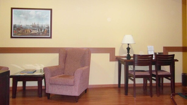 Um quarto com uma cadeira de madeira, uma mesinha com abajur e um quadro pendurado na parede.