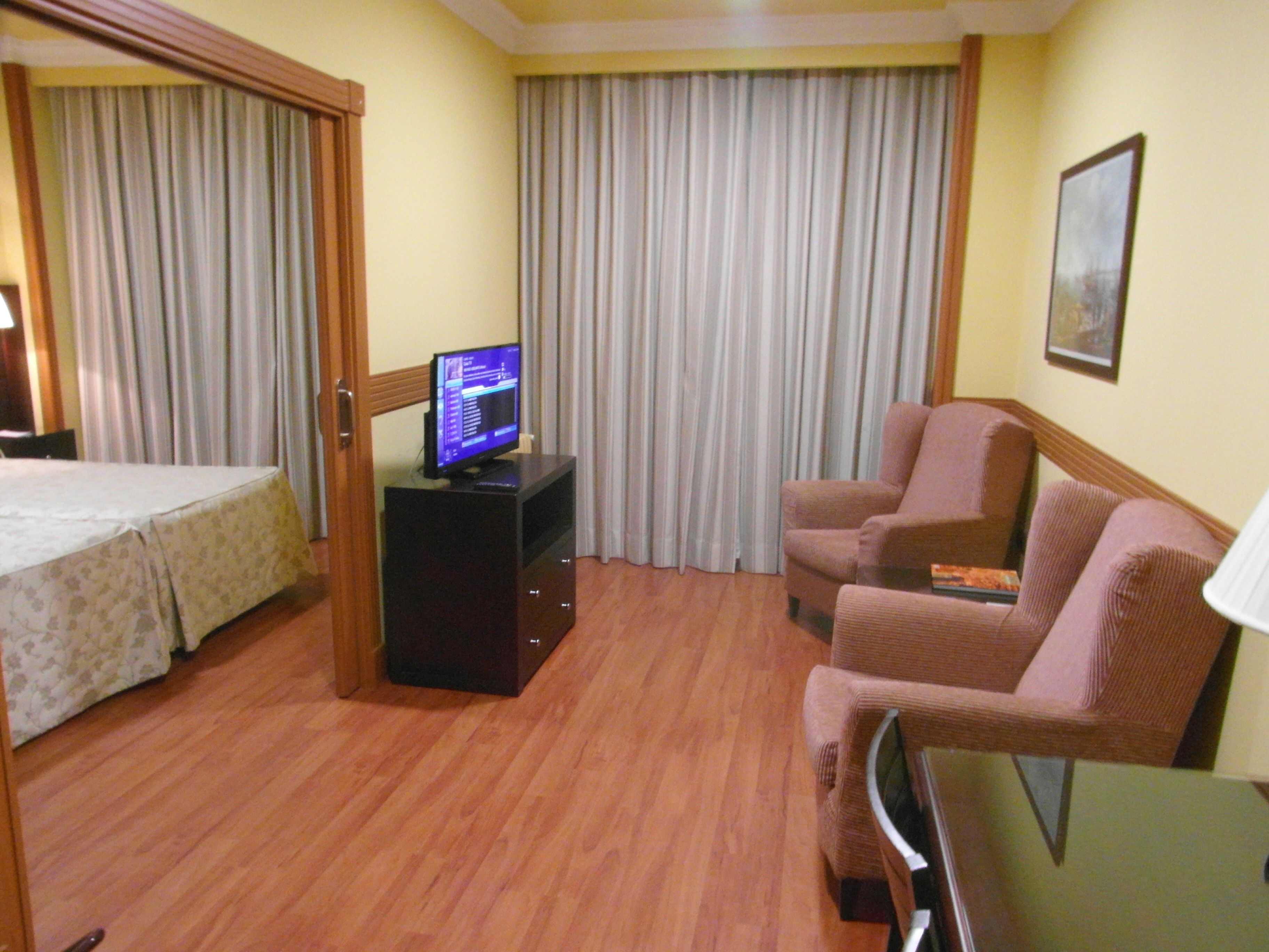 A hotel room with a bed, a TV on a stand, a seating area with two chairs, and a large window with curtains.