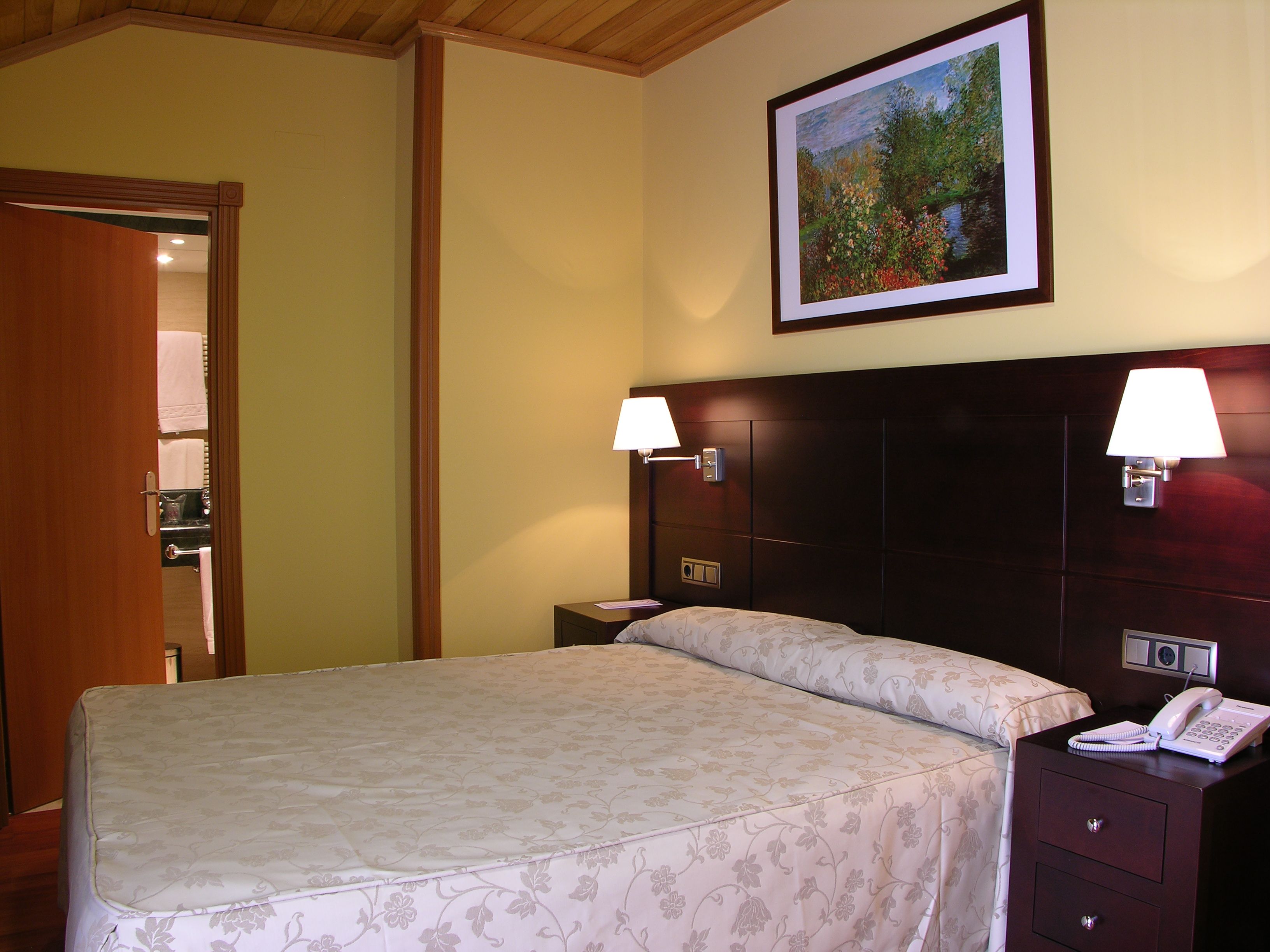 A hotel room with a bed, two bedside lamps, a nightstand with a phone, and a painting on the wall.