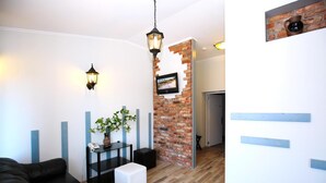 A modern living room with a black leather sofa, a white ottoman, a glass coffee table, and a brick accent wall with a mounted TV. - Apart Hotel TOMO (Riga)