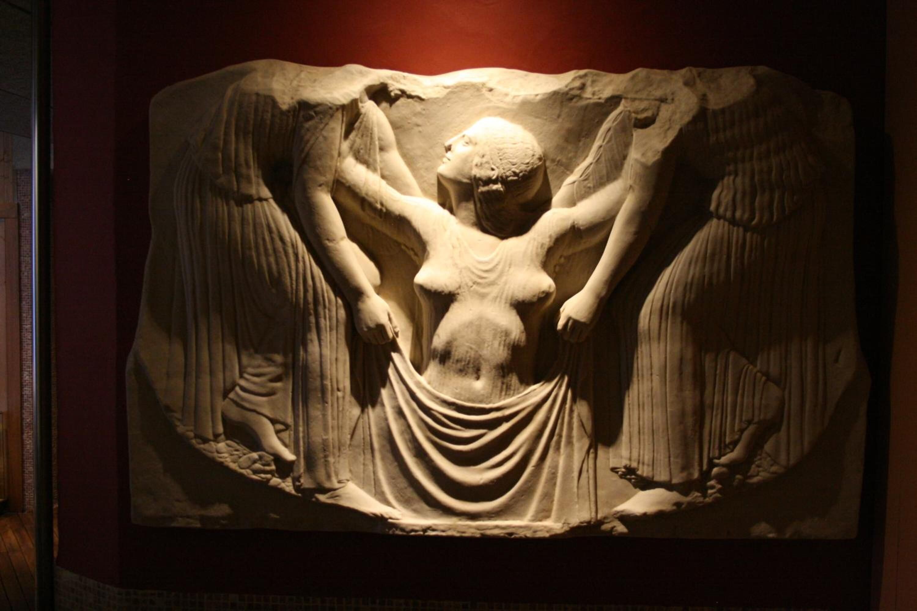 Una escultura de mármol con una figura central rodeada de alas, sobre un fondo oscuro.