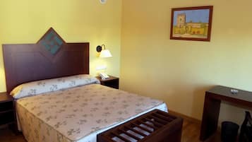 A hotel room with a bed, bedside table with a lamp, a desk, and a trash bin.