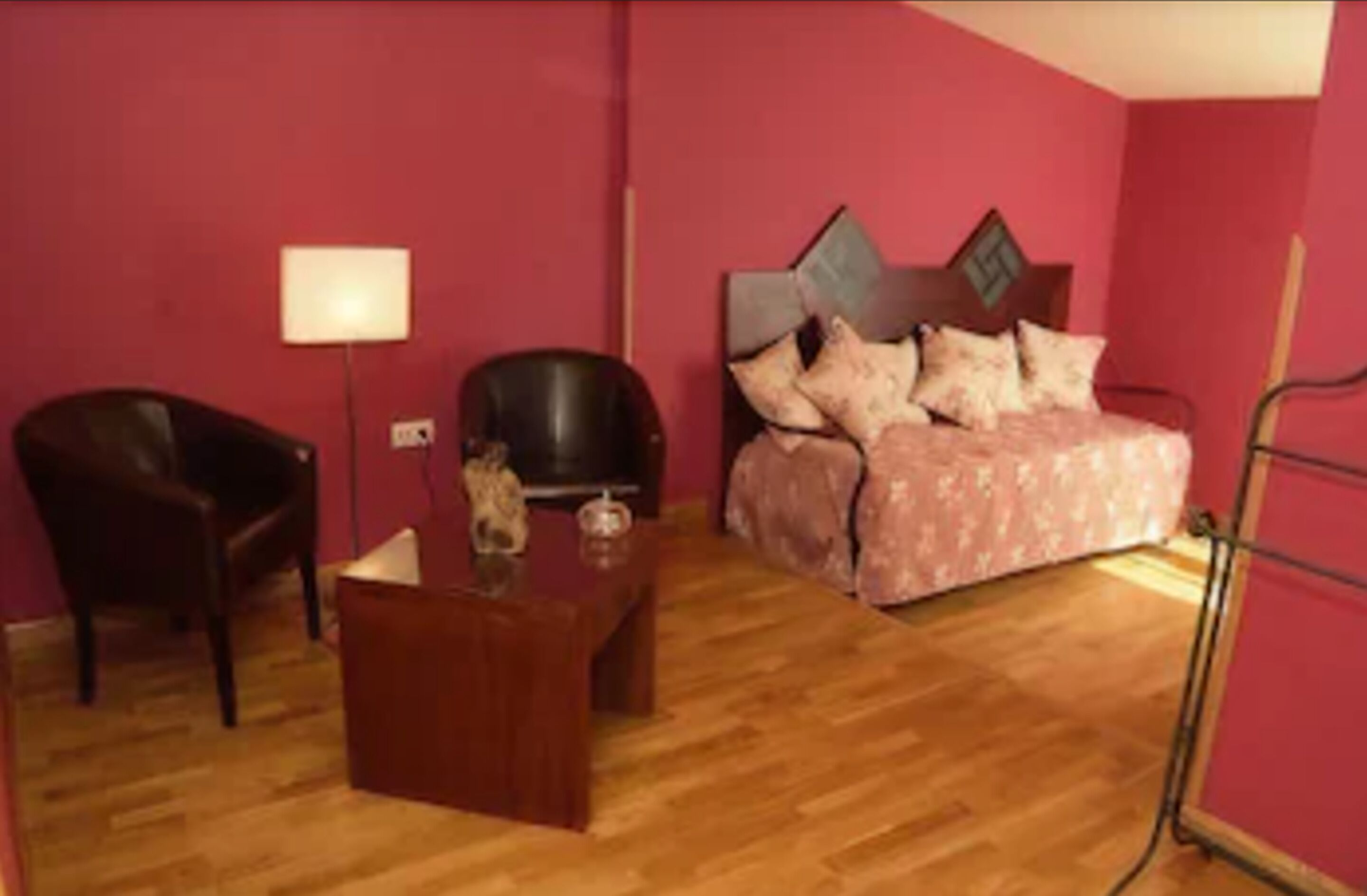 Une pièce avec un plancher en bois, un lit recouvert d’une couvre-lit à motifs floraux, un fauteuil en cuir noir, une petite table en bois et une lampe fixée au mur.