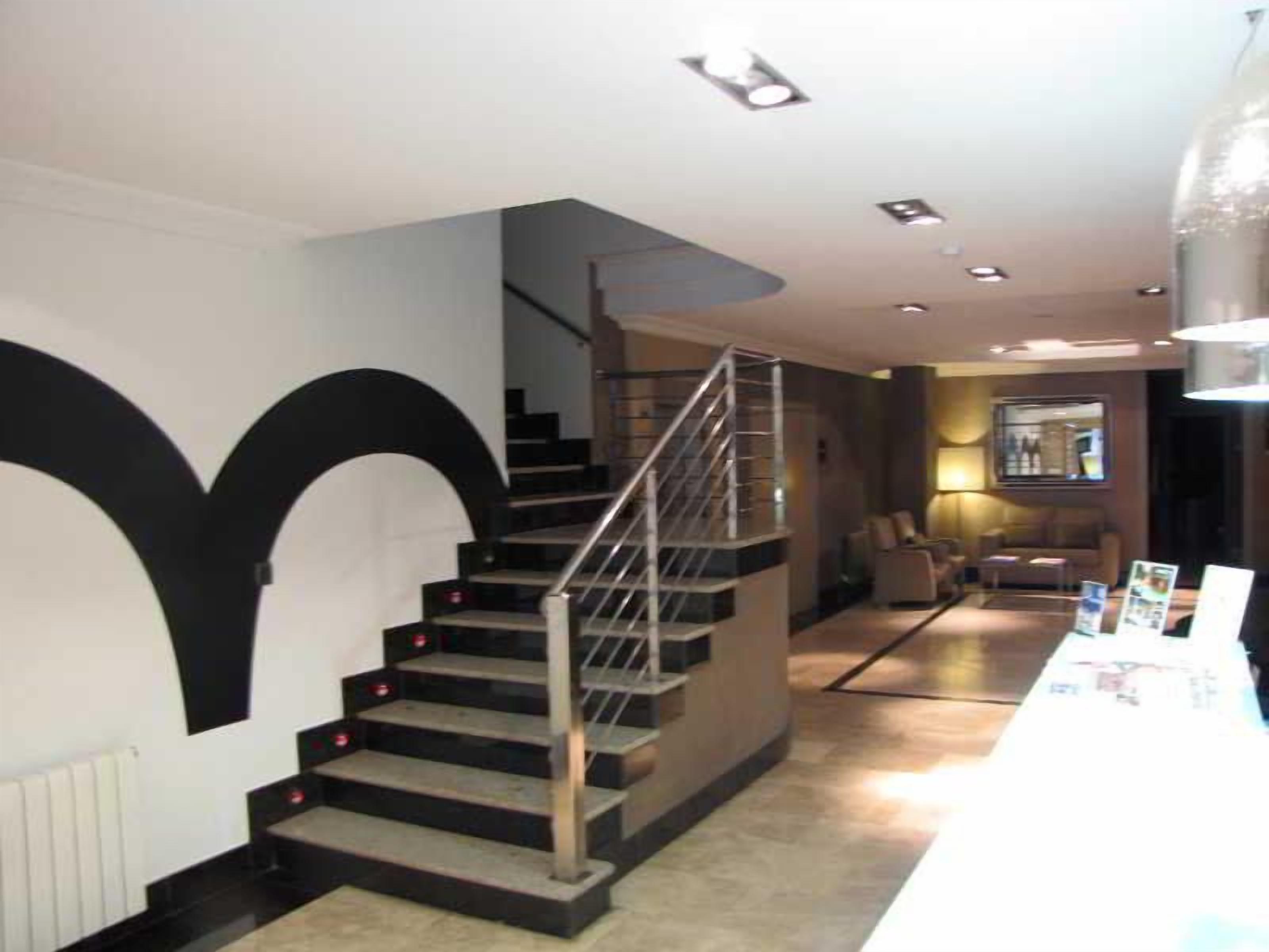 Una escalera con barandilla metálica que conduce a un piso superior, una pared blanca con un diseño negro grande y un mostrador de recepción con folletos.