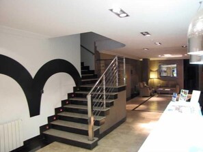 A staircase with a metal railing leading to an upper floor, a white wall with a large black design, and a reception desk with brochures. - Room Hotel (Pontevedra)