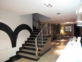 Una escalera con barandilla metálica que conduce a un piso superior, una pared blanca con un diseño negro grande y un mostrador de recepción con folletos.