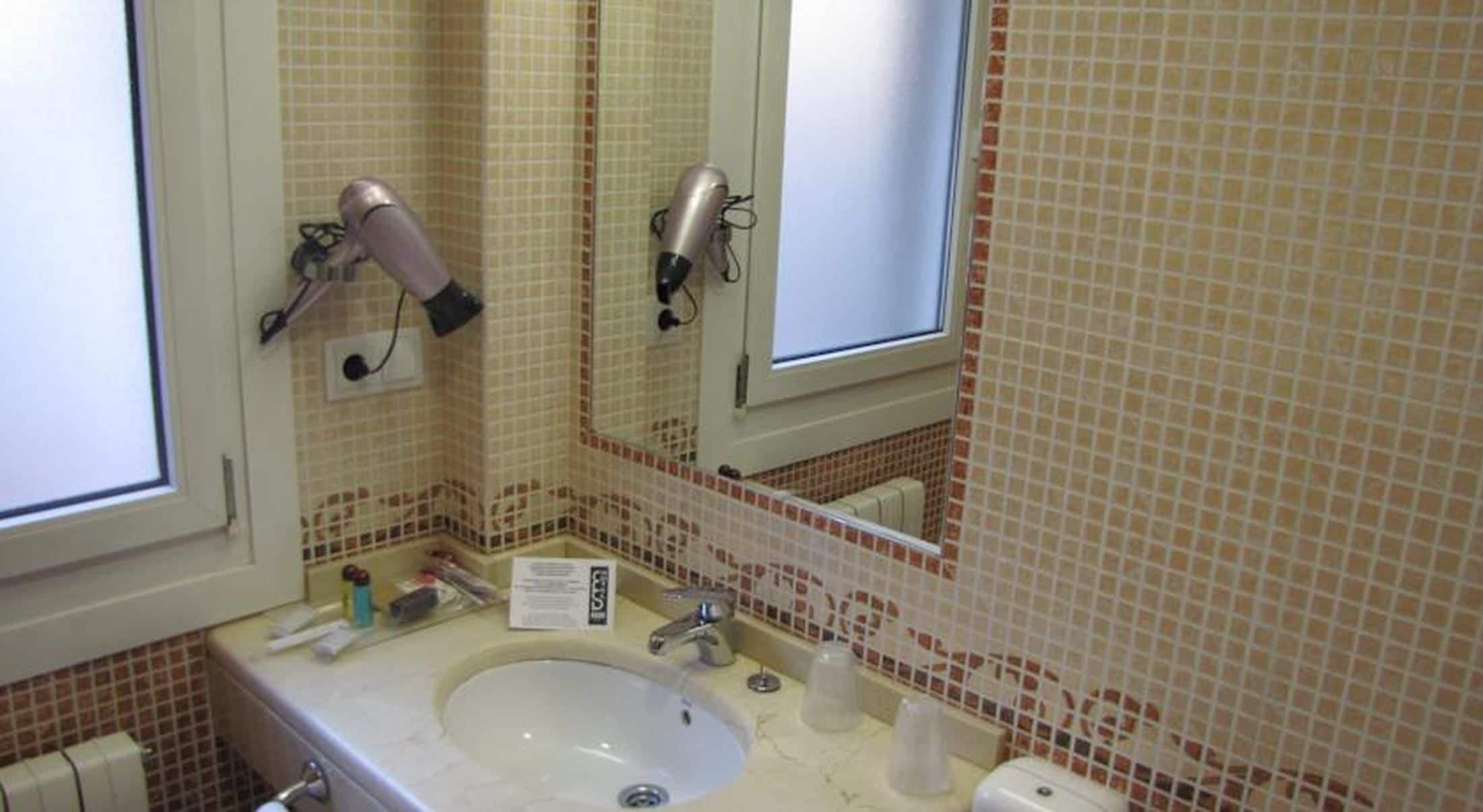 Un baño con un lavabo, dos secadores de pelo fijados a la pared, un espejo y una ventana.