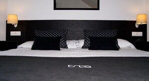 Una cama con ropa de cama blanco y negro, dos almohadas negras con diseño de diamantes y dos lámparas de pared a cada lado.