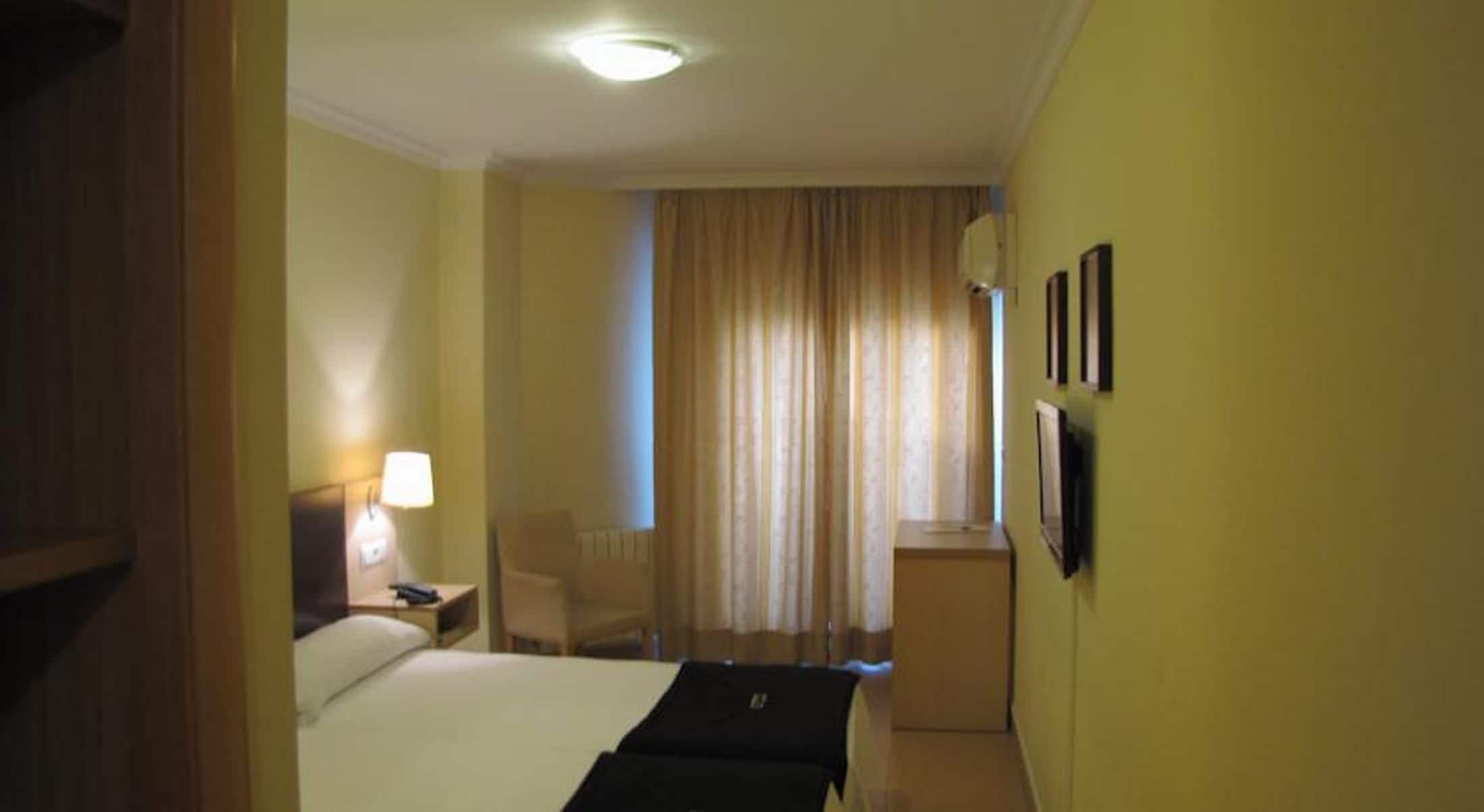 A hotel room with a bed, a desk, a chair, a lamp, and a window with curtains.