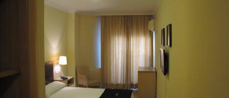 A hotel room with a bed, a desk, a chair, a lamp, and a window with curtains.