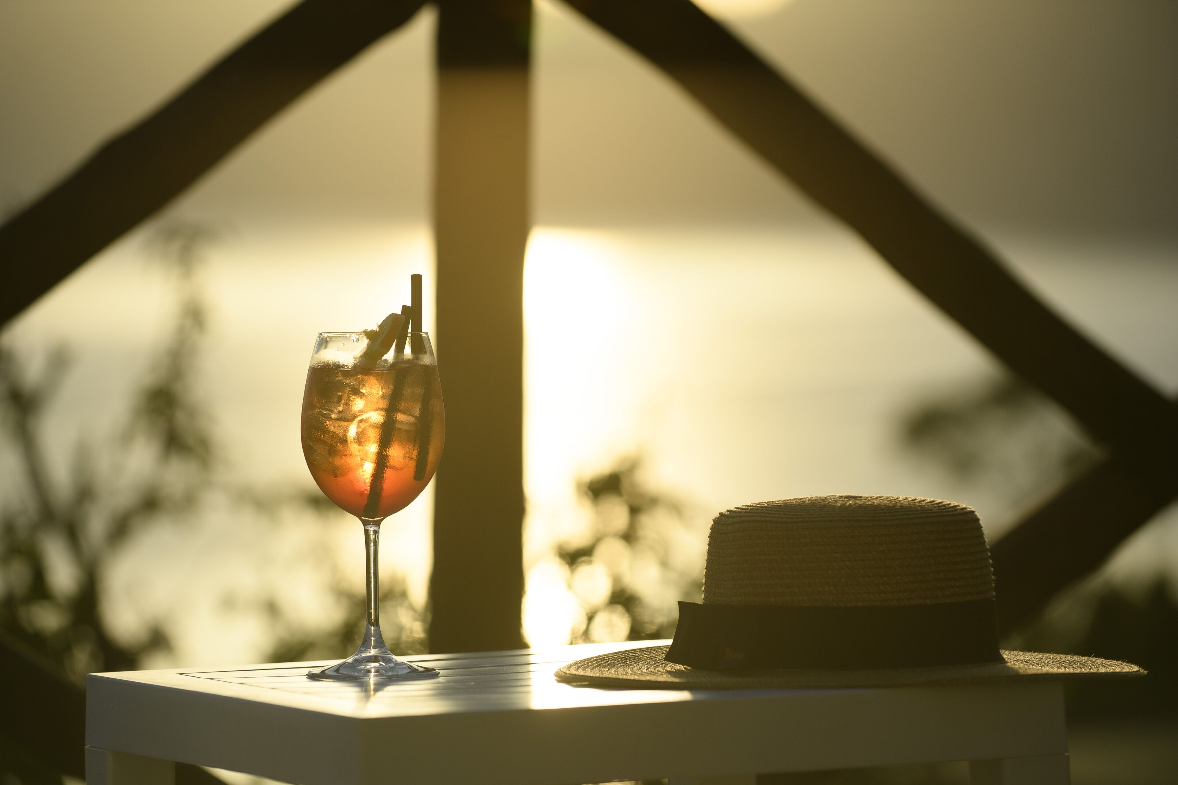 Un verre rempli de boisson sur une table, avec un chapeau à côté, le tout sur fond de coucher de soleil.