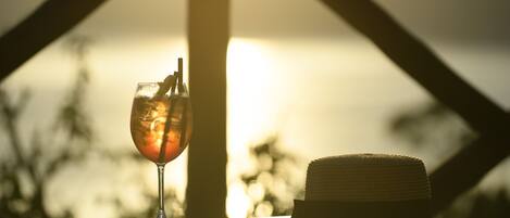 Een drankje in een glas op een tafel, met een hoed ernaast, voor een zonsondergang.