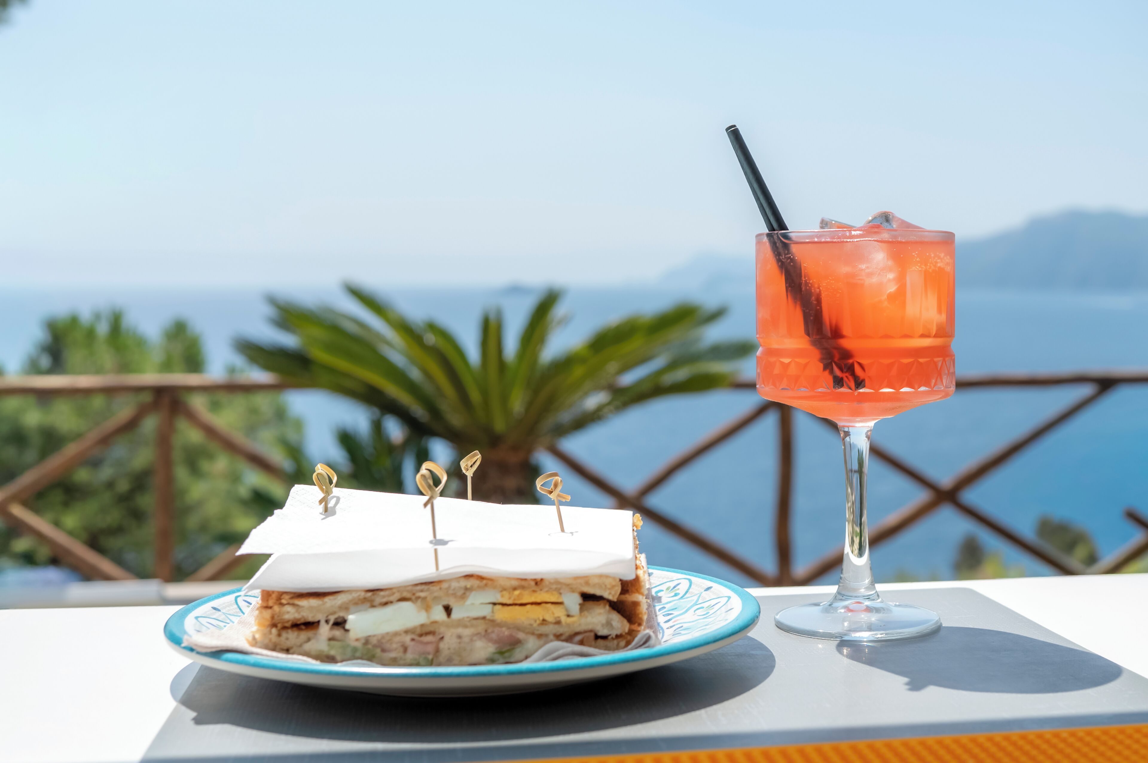 Un sándwich con palillos sobre un plato, un vaso de bebida y vista al mar.