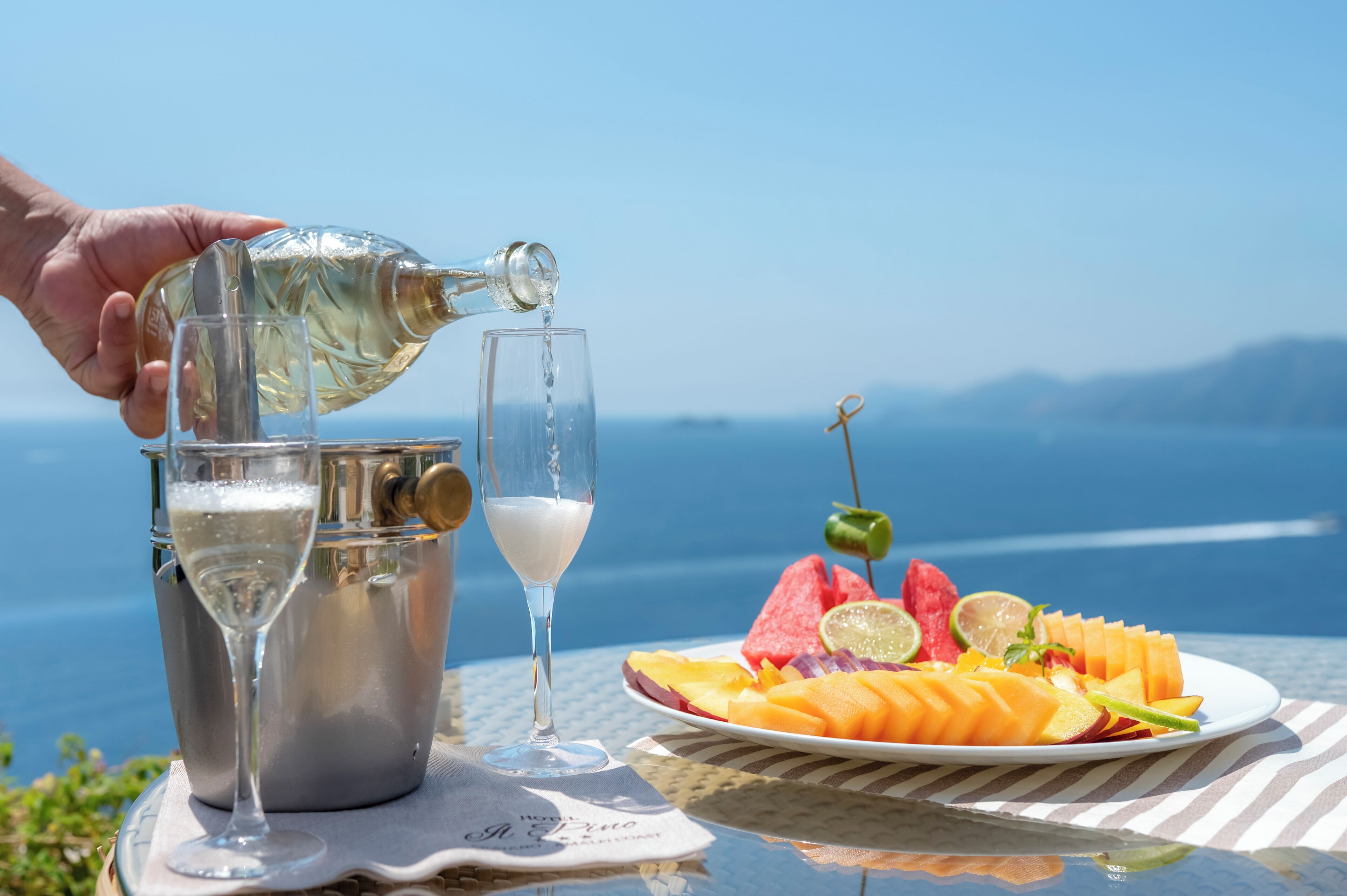 Een hand giet een drankje in een glas, een waterfles en een schaal fruit op een tafel met een prachtig uitzicht.