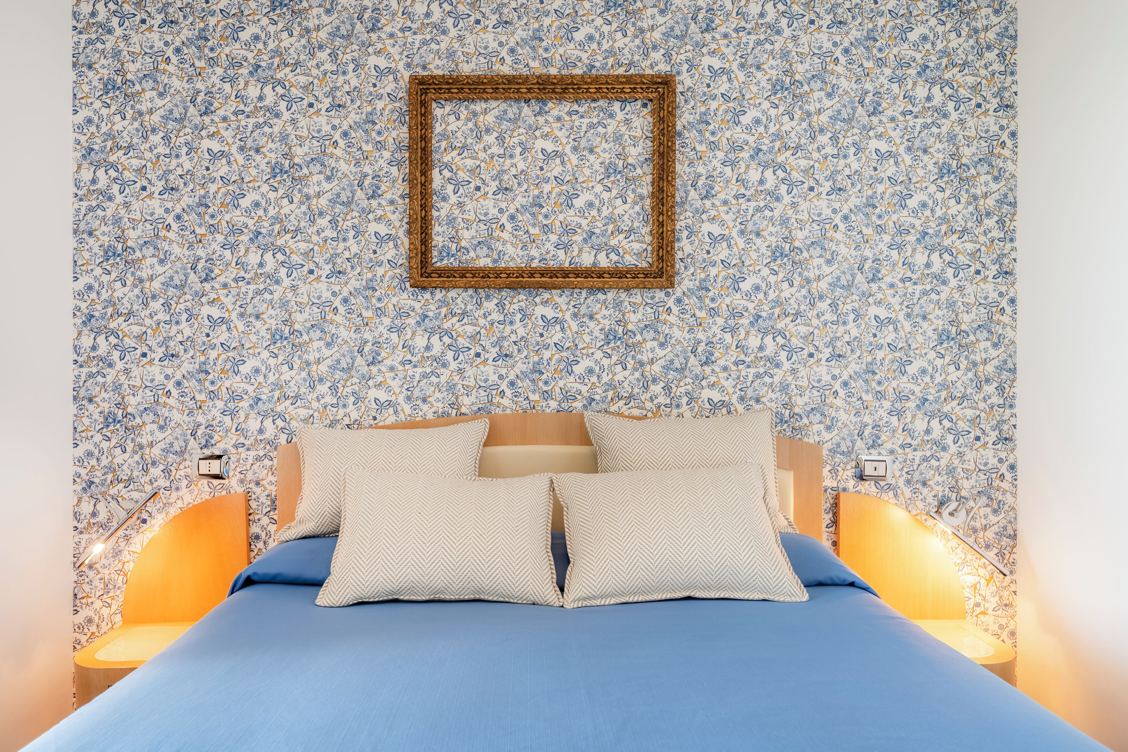A bedroom with a floral-patterned wall, a bed with blue bedding, and two bedside lamps.