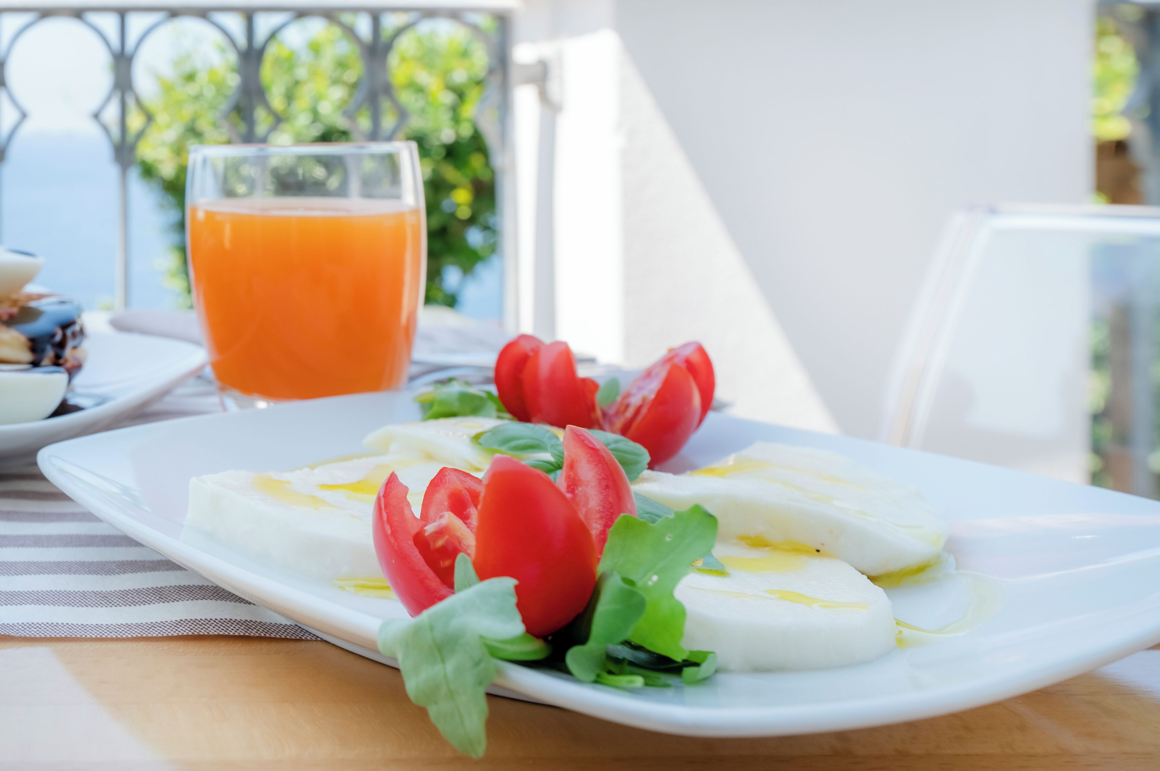 Ein Teller frischer Mozzarella mit Tomaten und Basilikum, ein Glas Orangensaft und eine gestreifte Serviette auf einem Holztisch.