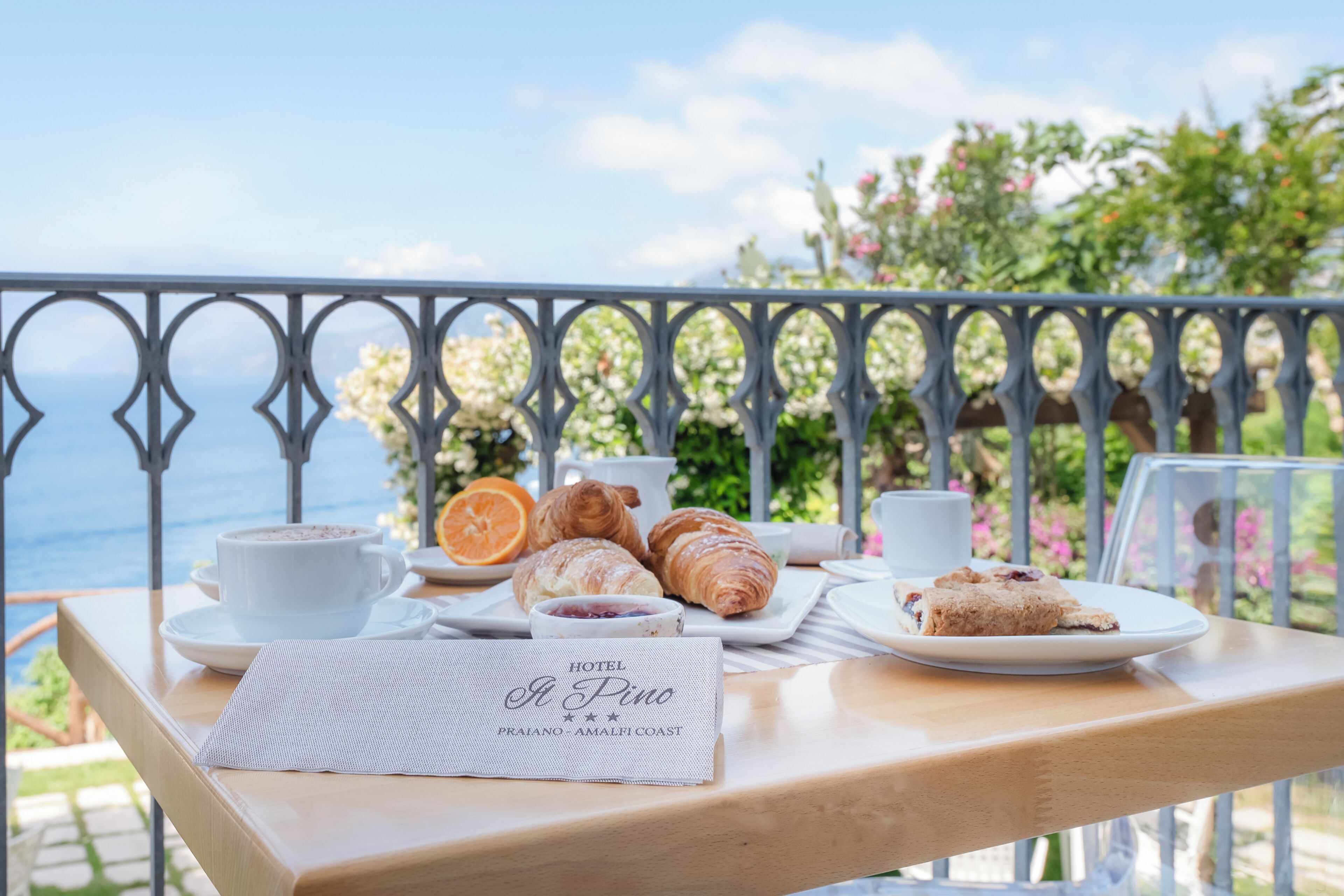 Een tafel met croissants, gebak en een kop koffie op een balkon met uitzicht op zee.