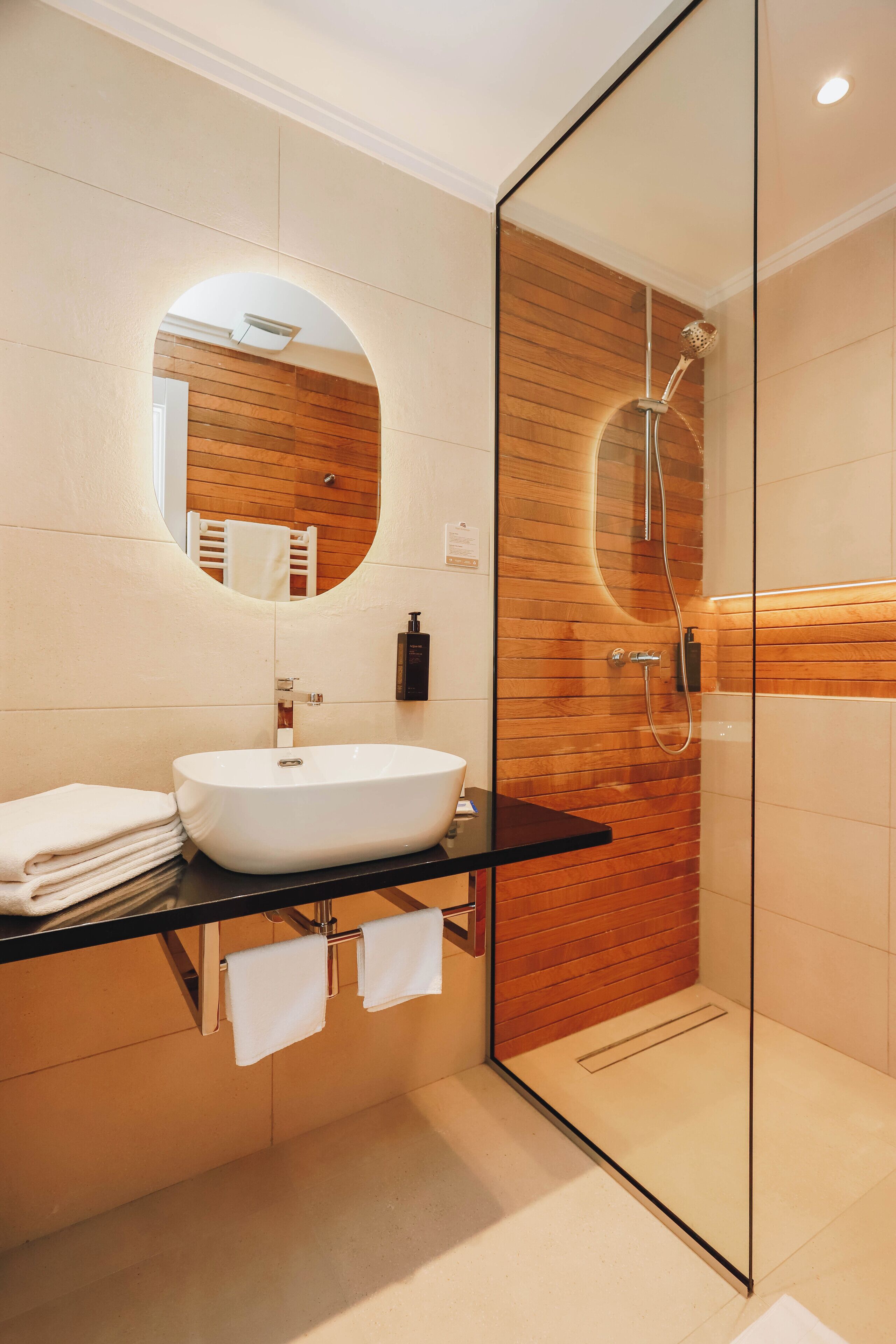Un baño moderno con ducha de vidrio, un espejo redondo y un lavamanos blanco.