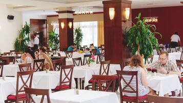A restaurant with multiple tables and chairs, indoor plants, and a modern chandelier.