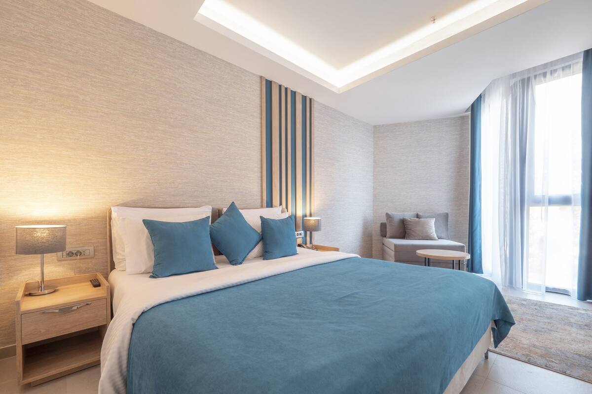 A modern hotel room with a large bed, bedside tables, a sofa, and a view of the outdoors.