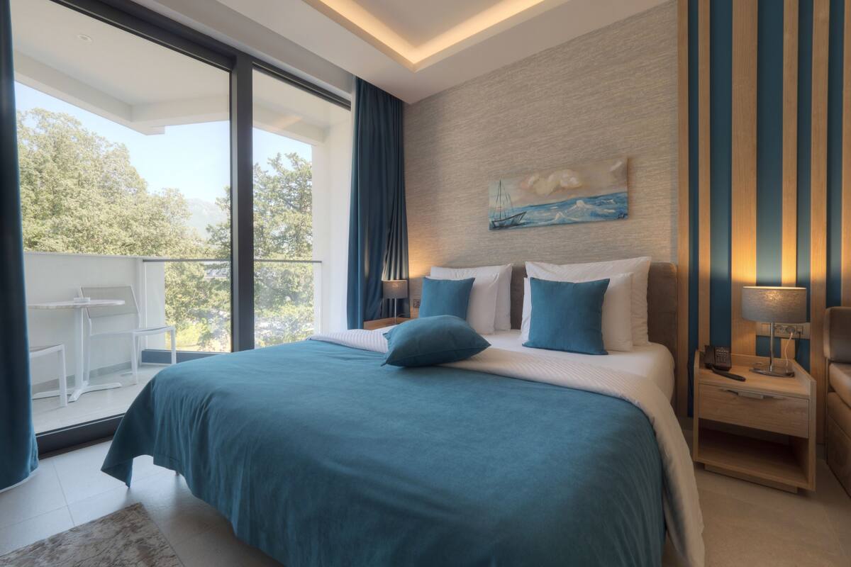 A modern hotel room with a large bed, a balcony with a table and chairs, and a view of trees.