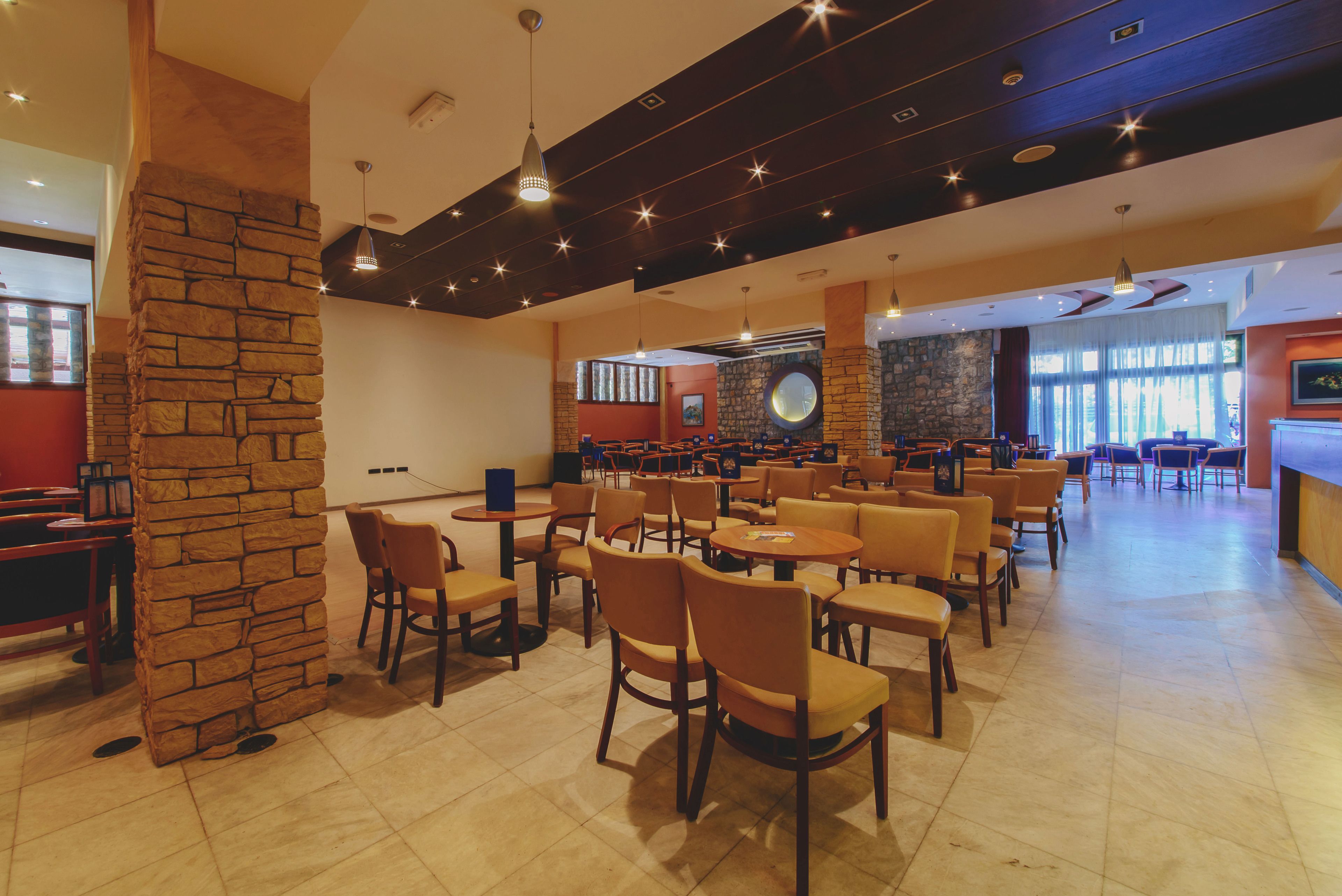 A spacious restaurant with multiple tables and chairs, a stone pillar, and a stone wall with a circular window.