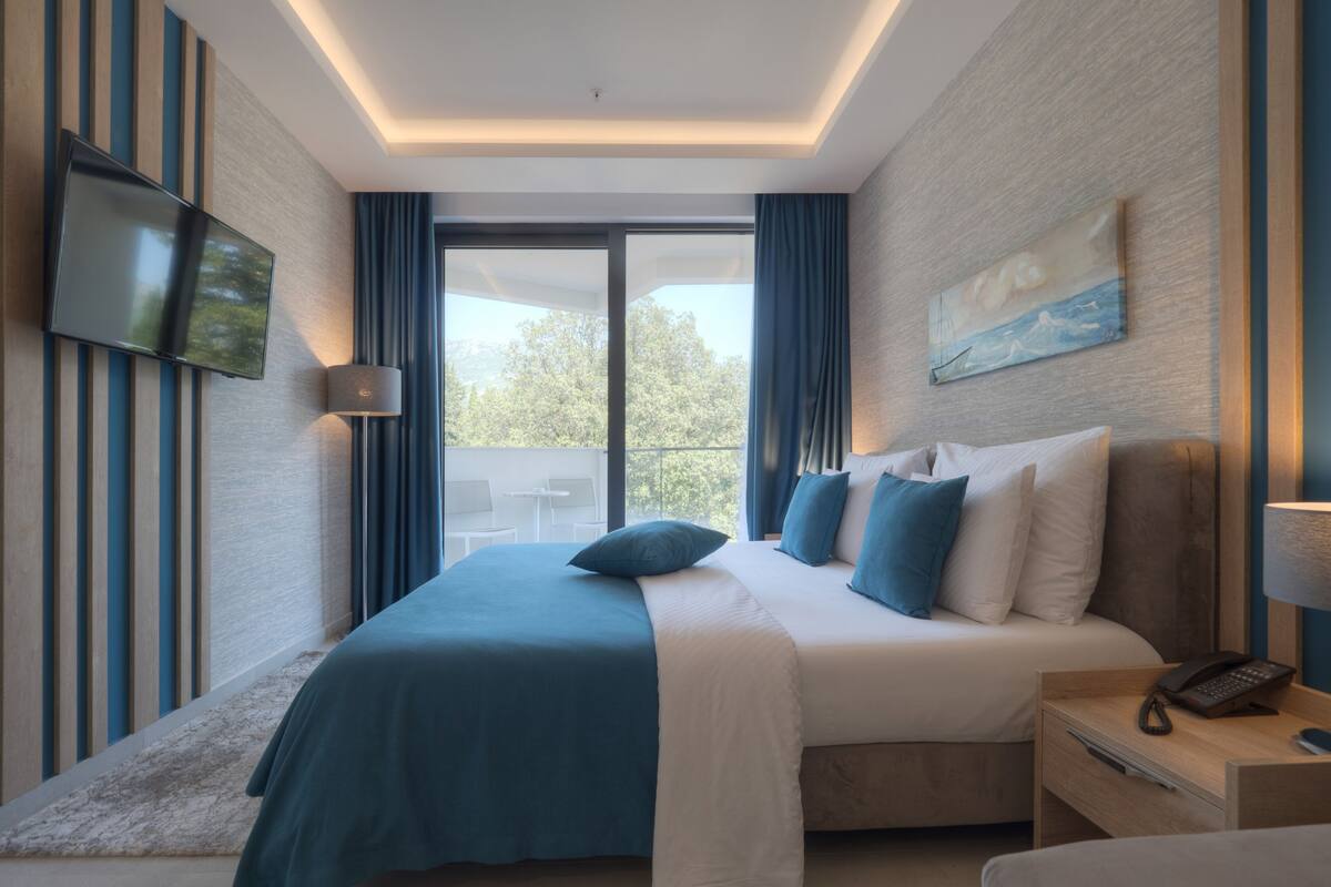 A modern hotel room with a large bed, a flat-screen TV mounted on the wall, a balcony with a table and chairs, and a painting on the wall.