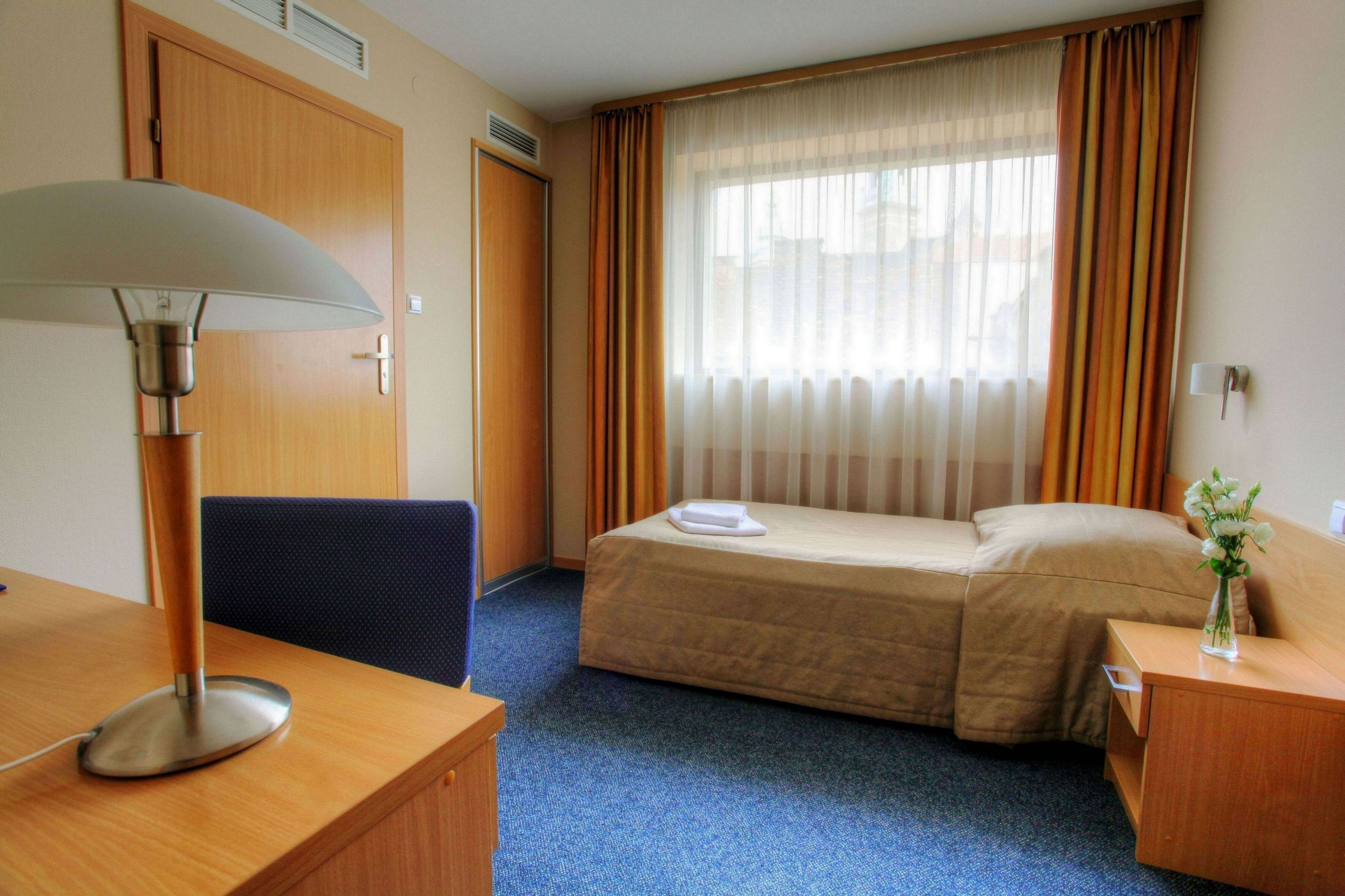 Una camera d'albergo con un letto, una scrivania con una lampada, una sedia, un comodino con un vaso e una finestra con tende.