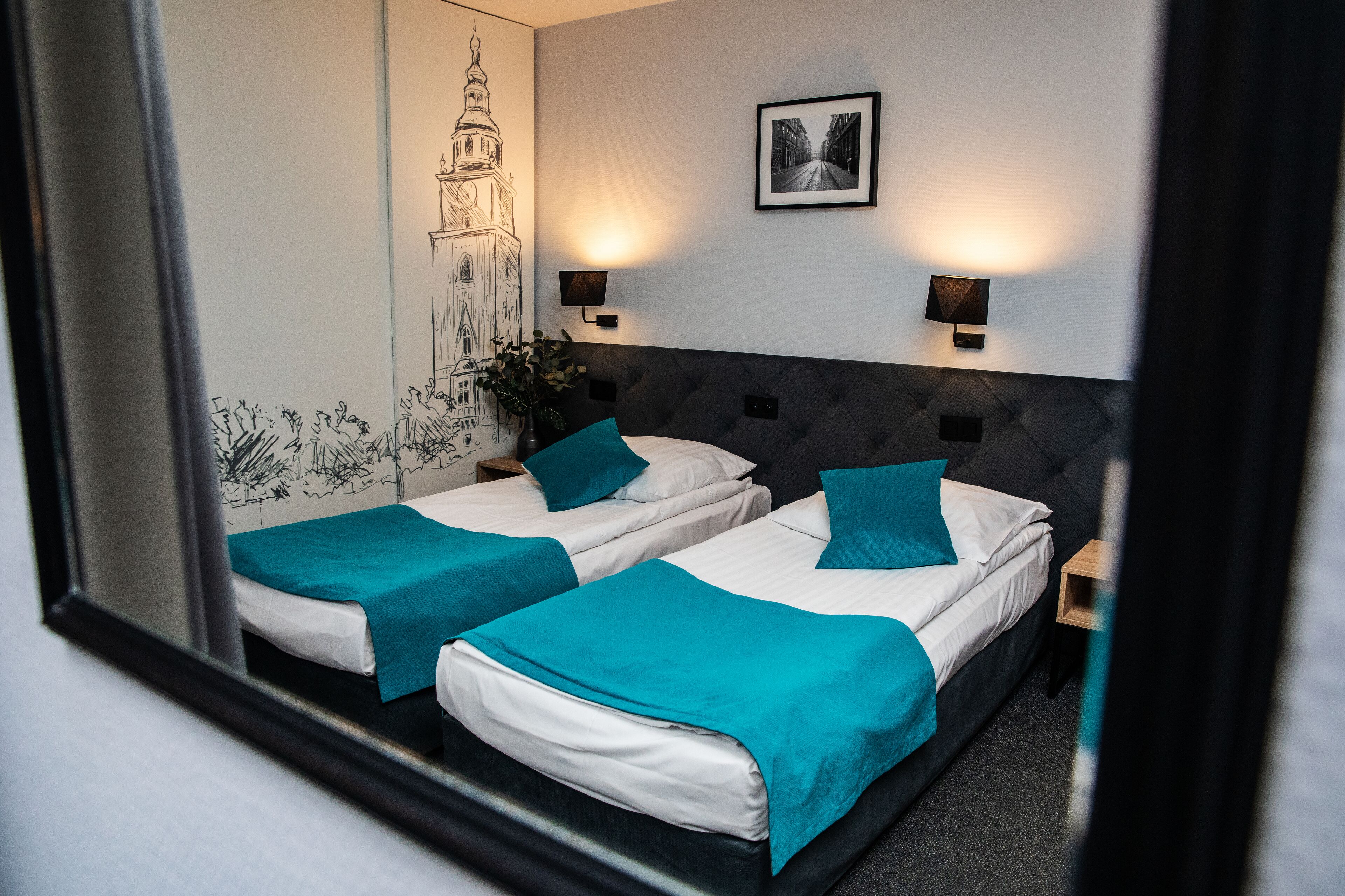 Habitación de hotel con dos camas, ropa de cama color turquesa, un espejo grande, un mural en la pared y dos lámparas fijadas a la pared.