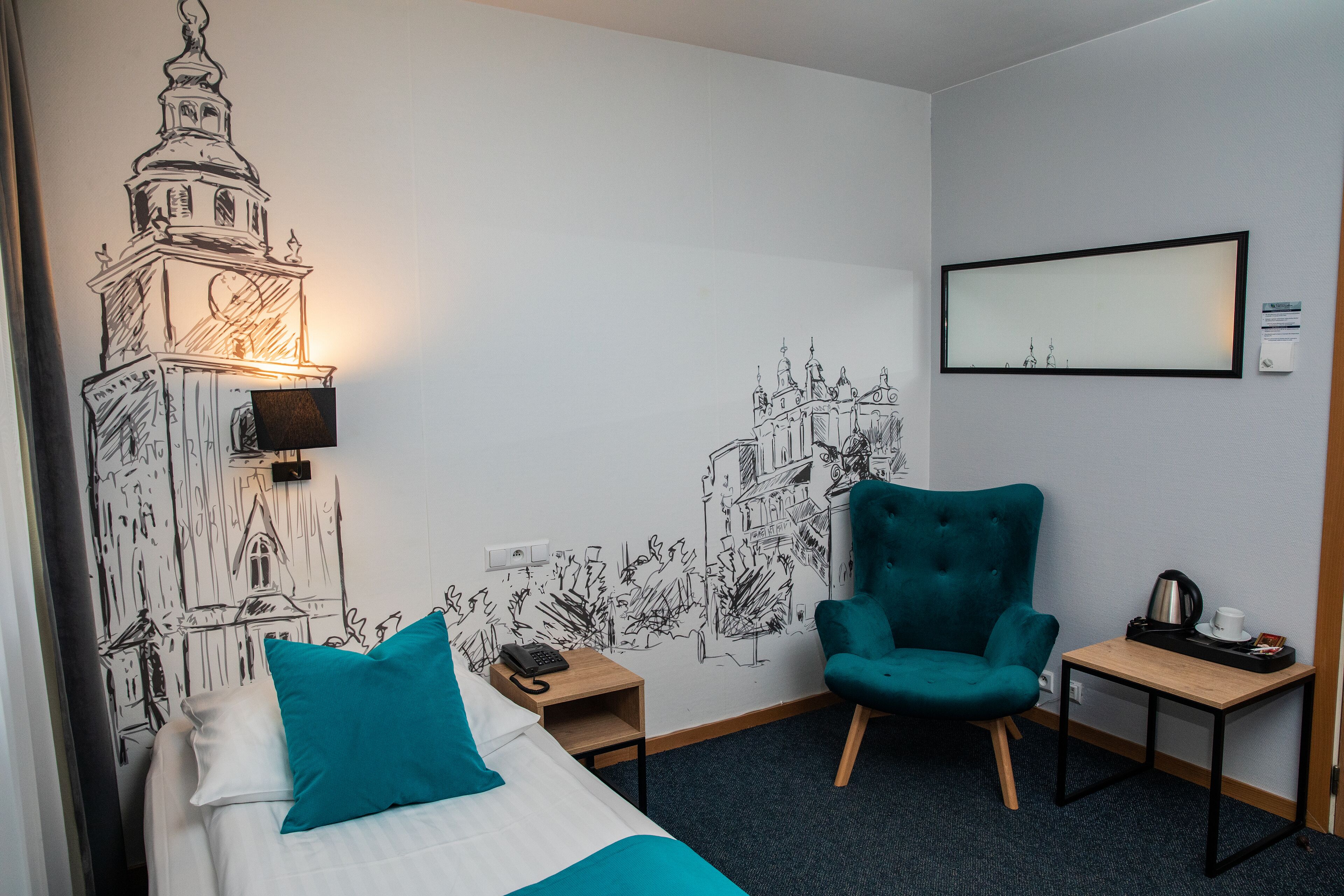 A hotel room with a bed, a teal armchair, a small table with a kettle, and a wall mural of a building.