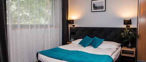 Una cama bien hecha con una manta color turquesa, dos lámparas de noche, un cuadro en la pared y una ventana con cortinas traslúcidas.
