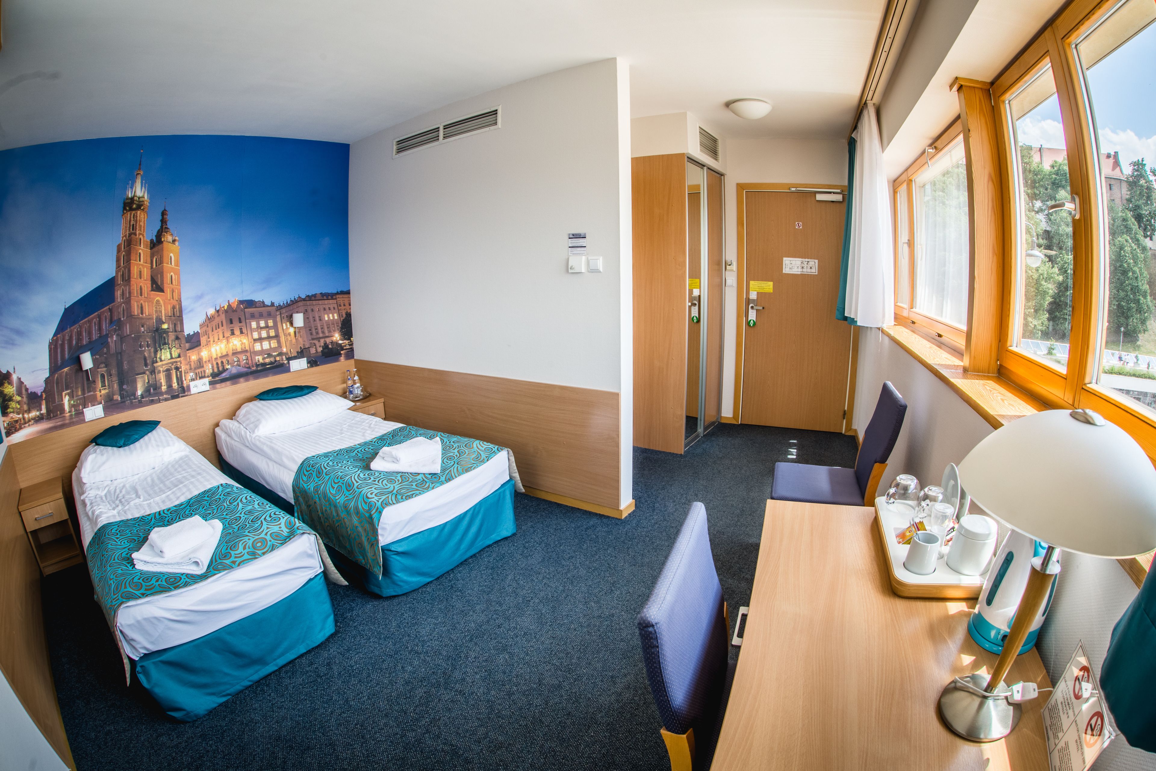 A hotel room with two beds, a desk with a lamp, a window with a view, and a wall with a cityscape mural.