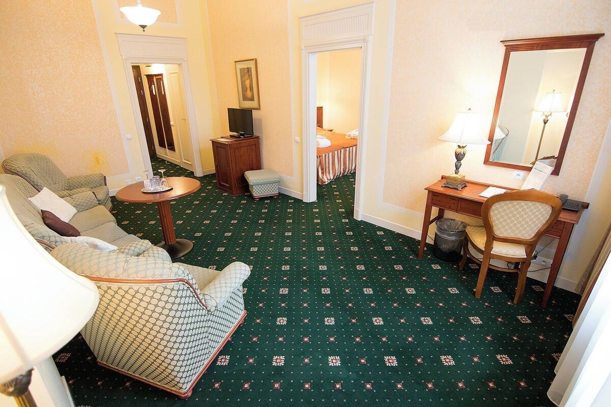 Habitación de hotel con alfombra verde estampada, un sofá, una silla, un escritorio con lámpara, un espejo y un televisor.