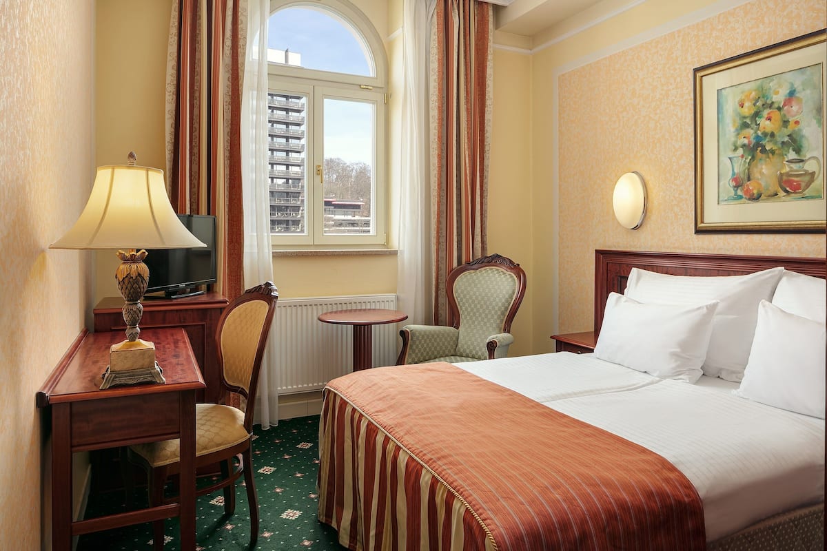 Habitación de hotel con cama, escritorio con lámpara, silla, mesita y ventana con cortinas.