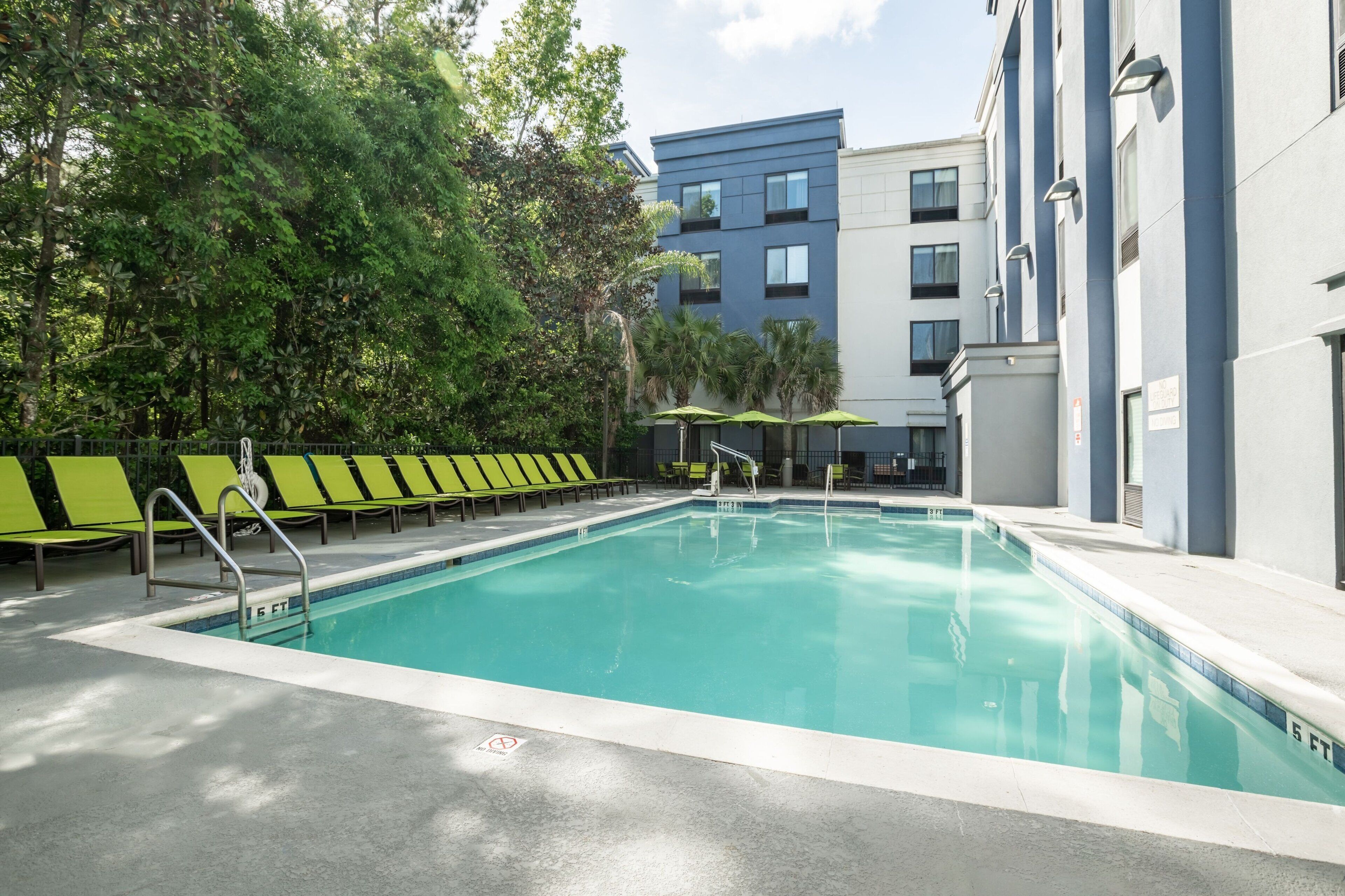 A swimming pool with green lounge chairs and umbrellas, surrounded by trees and a multi-story building.