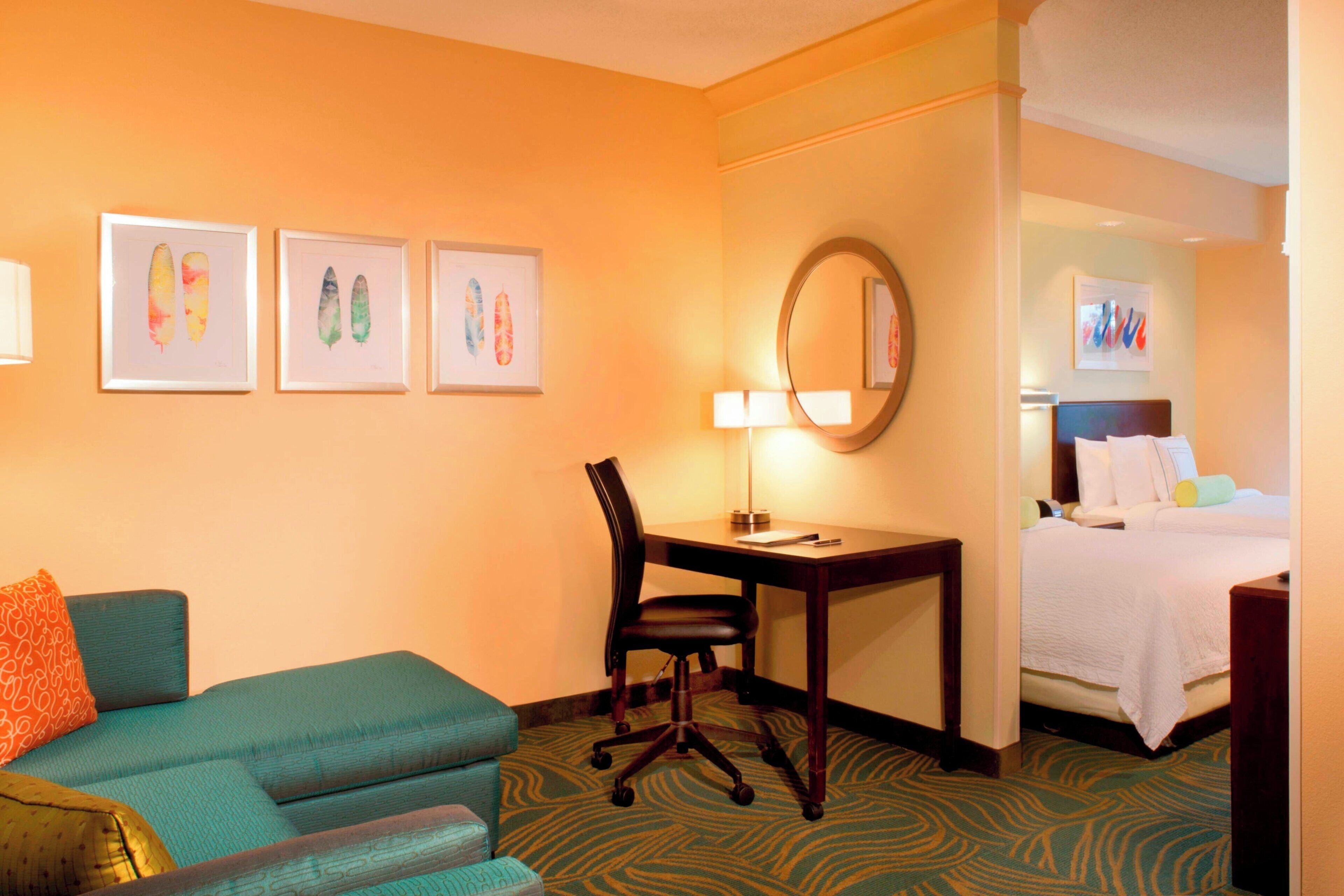 A hotel room with a teal sofa, a desk with a chair, a bed with white bedding, and three framed artworks on the wall.