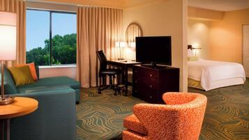 A hotel room with a bed, a desk with a television, a sofa, an armchair, and a view of trees through the window.