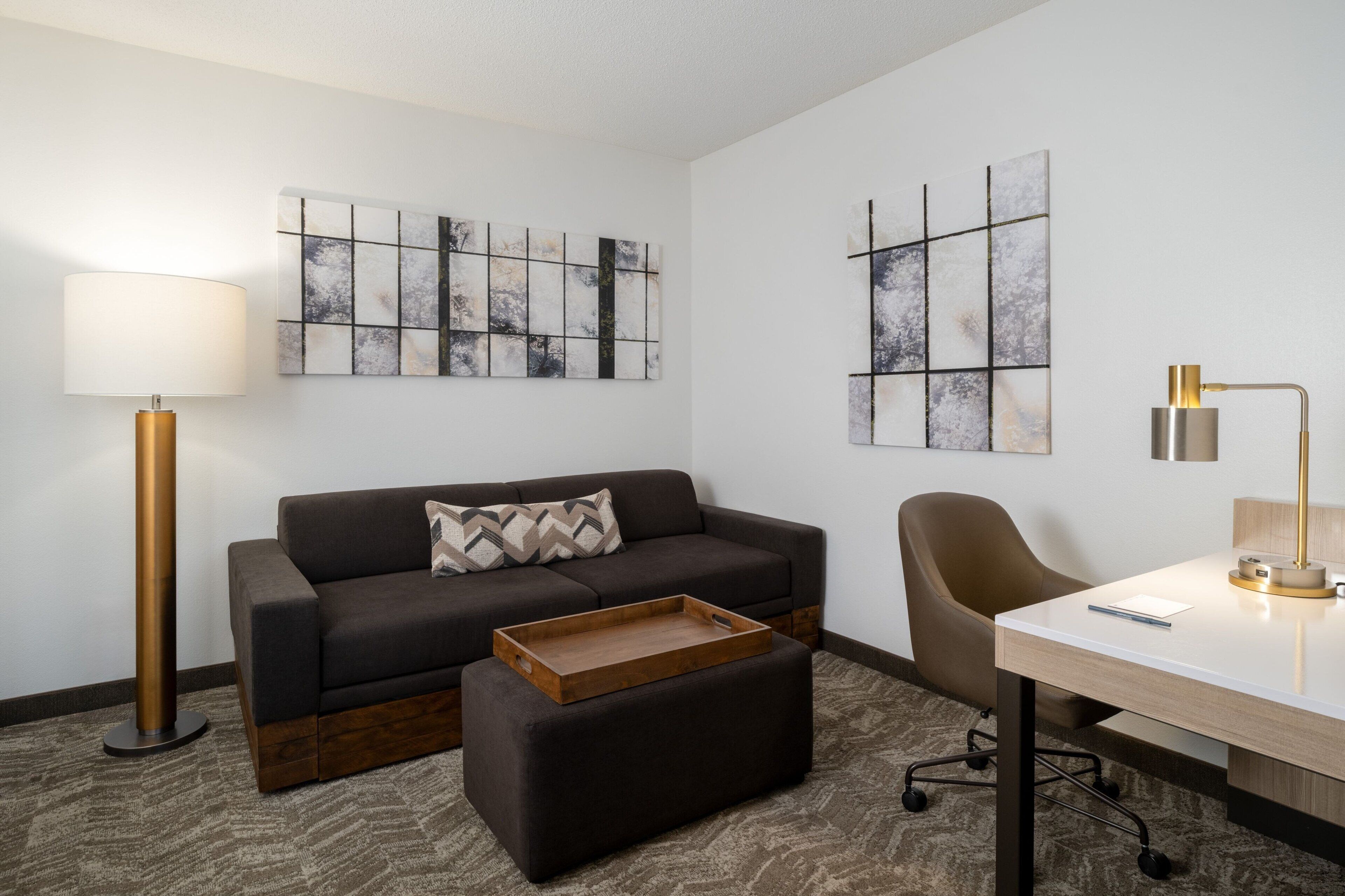 A modern room with a dark brown sofa, a wooden ottoman, a desk with a lamp, and abstract wall art.