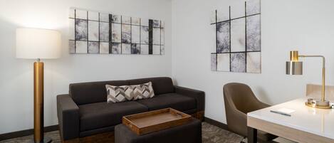 A modern room with a dark brown sofa, a wooden ottoman, a desk with a lamp, and abstract wall art.