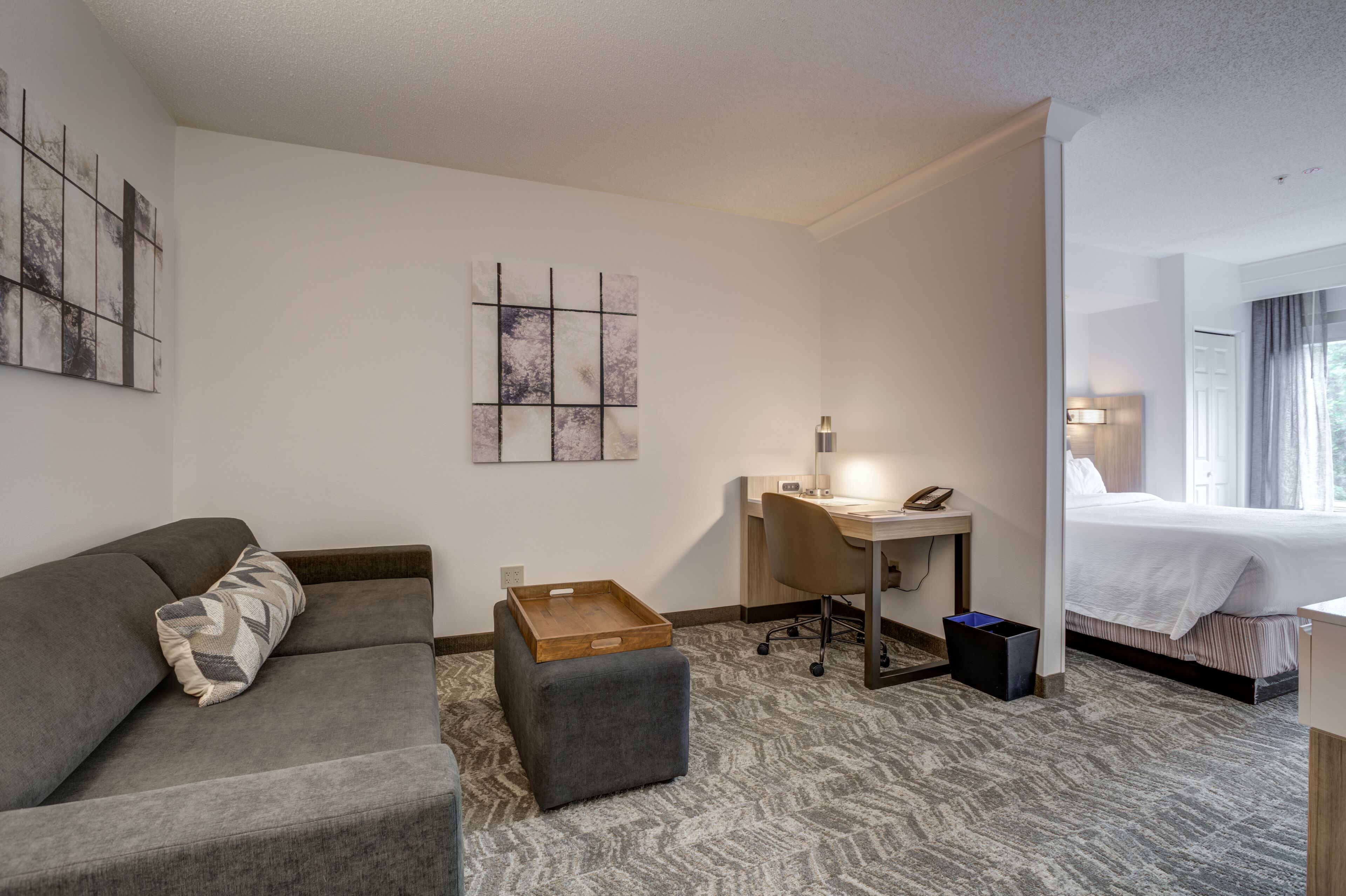 A hotel room with a gray sofa, a small desk with a lamp, a bed, and artwork on the walls.