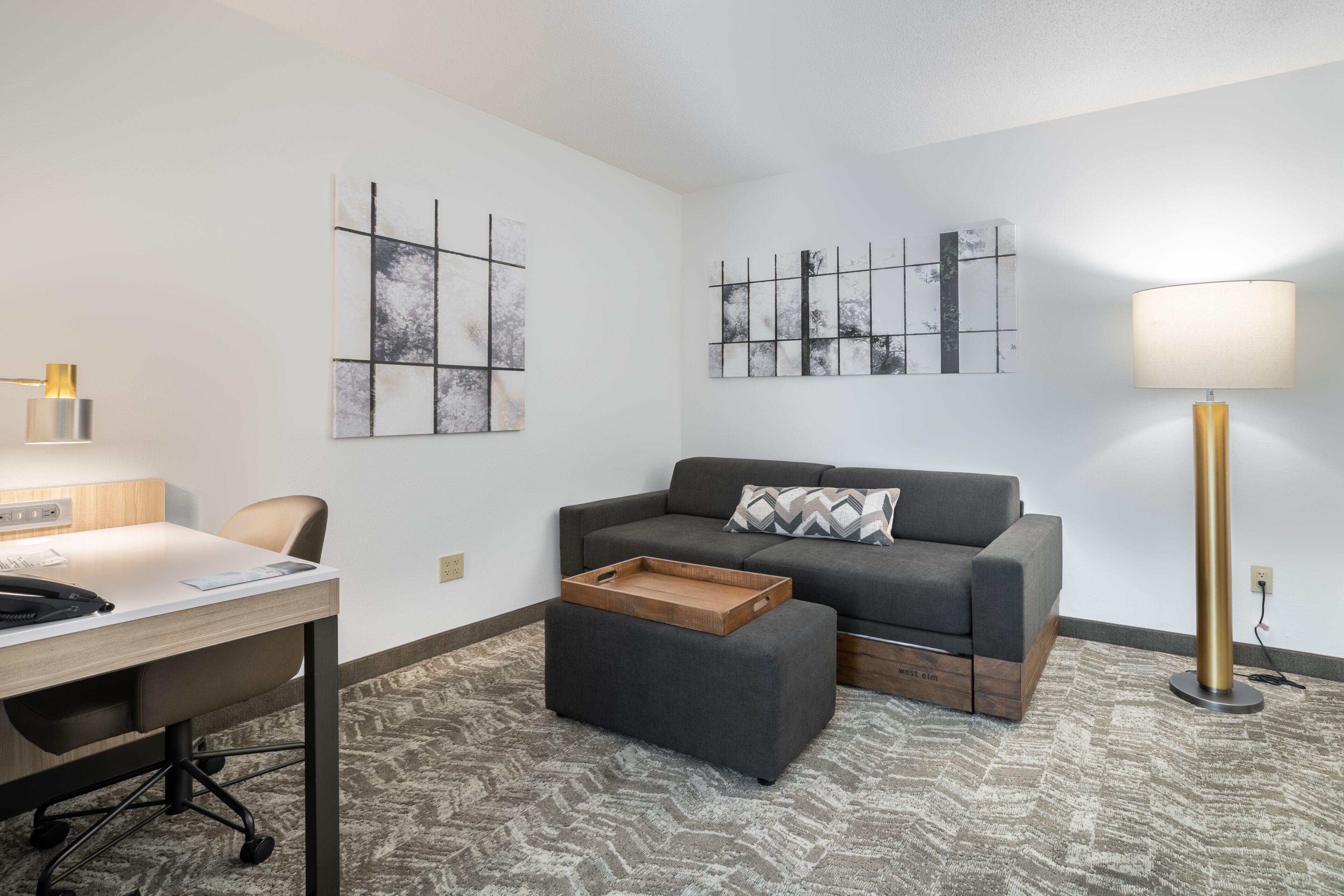 Een moderne hotelkamer met een grijze bank, een houten bijzettafeltje, een bureau met lamp en abstracte wandkunst.
