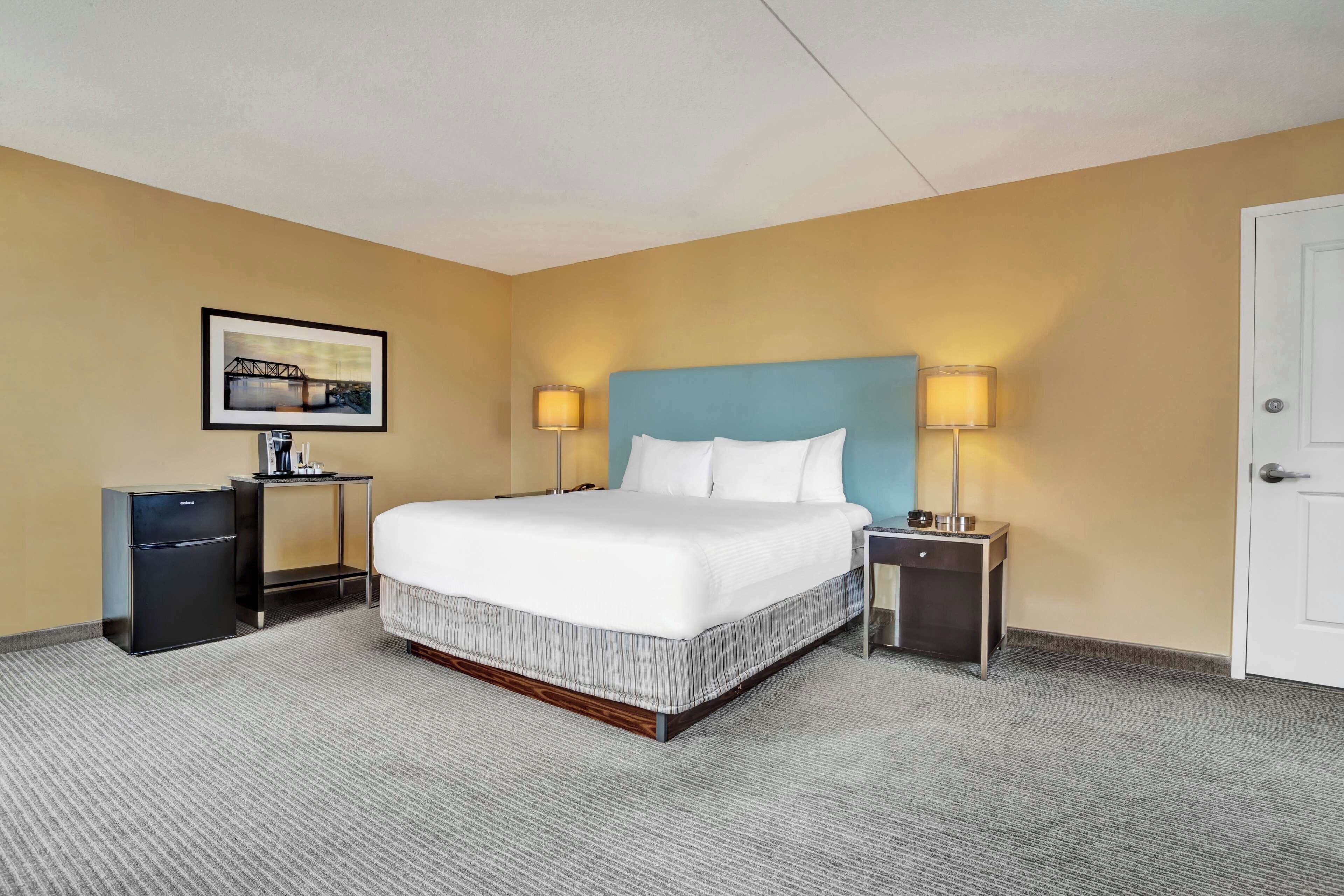 A hotel room with a bed, bedside tables, a refrigerator, a picture on the wall, and a door.