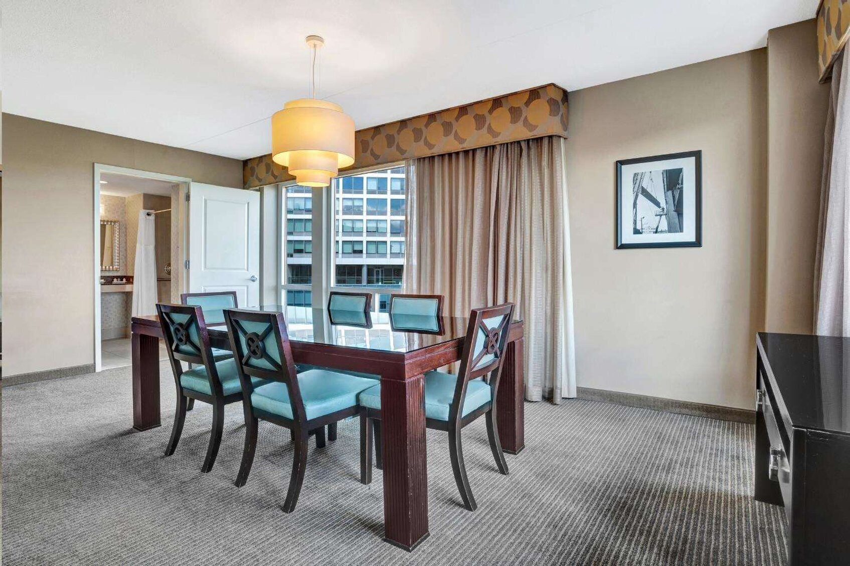 A hotel dining area with a round table, four chairs, a hanging light fixture, a window with curtains, and a framed picture on the wall.