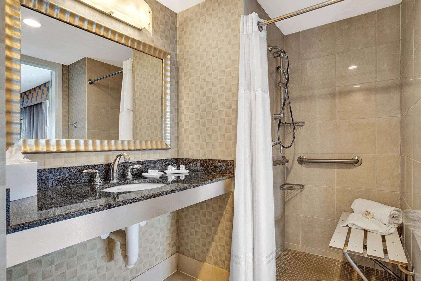 A modern bathroom with a granite countertop, a large mirror, a shower with a handheld showerhead, and a towel rack.