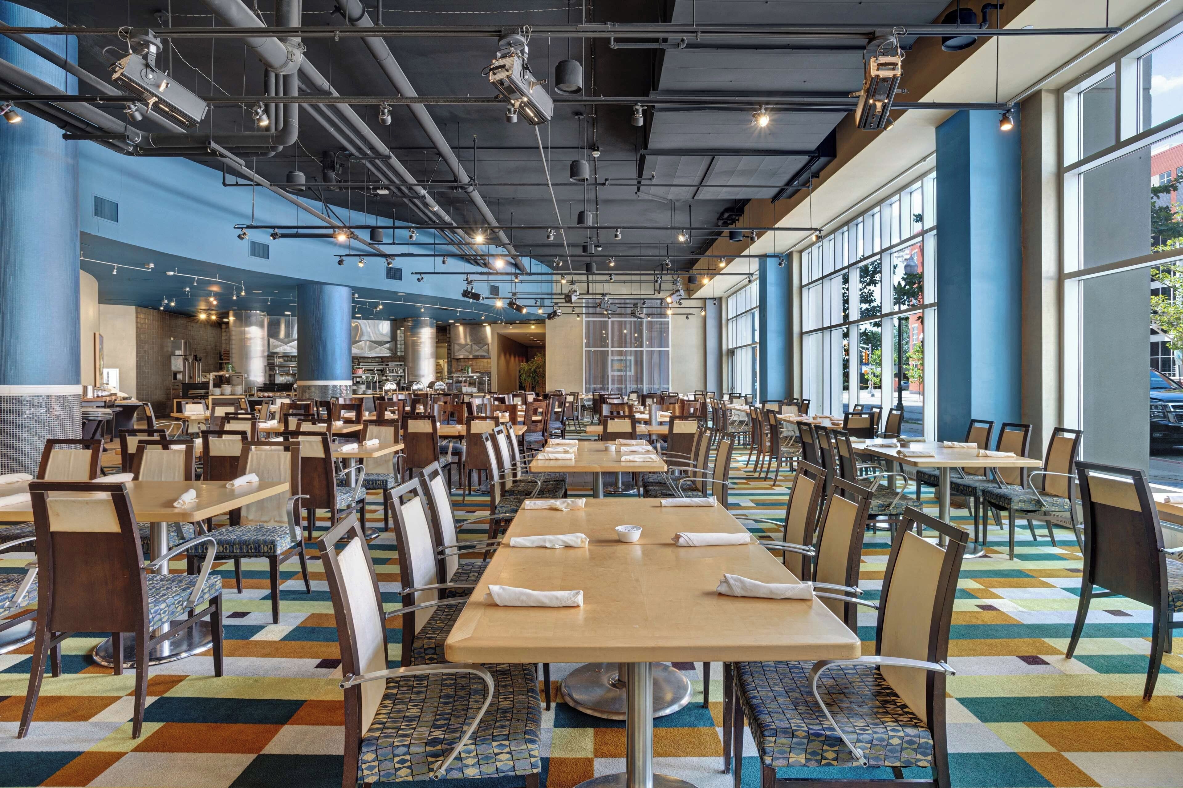 A spacious dining area with multiple tables and chairs, large windows, and a high ceiling with exposed pipes.