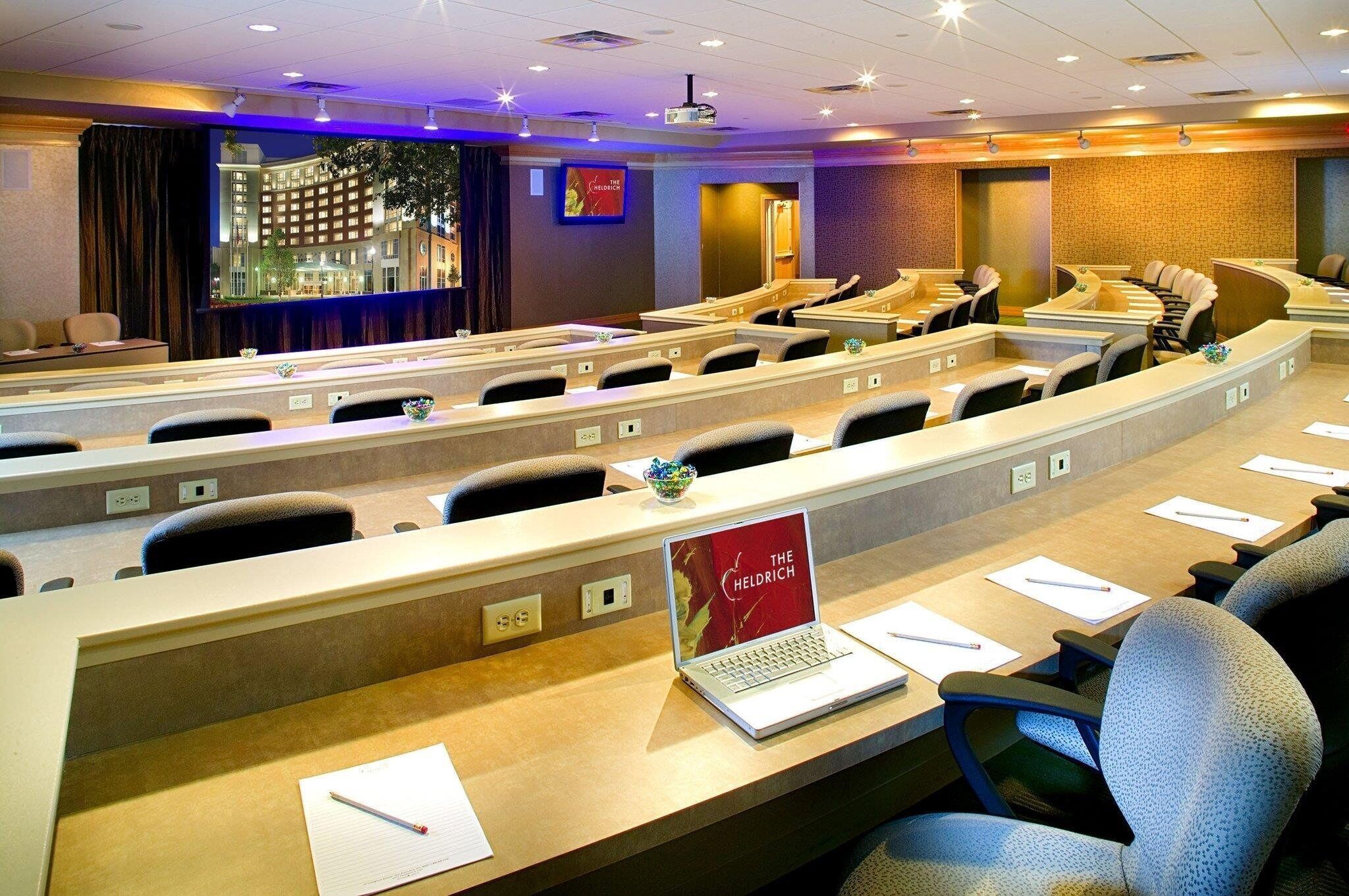 A conference room with rows of tables and chairs, a projector screen, and a laptop on one table.