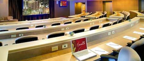 A conference room with rows of tables and chairs, a projector screen, and a laptop on one table.