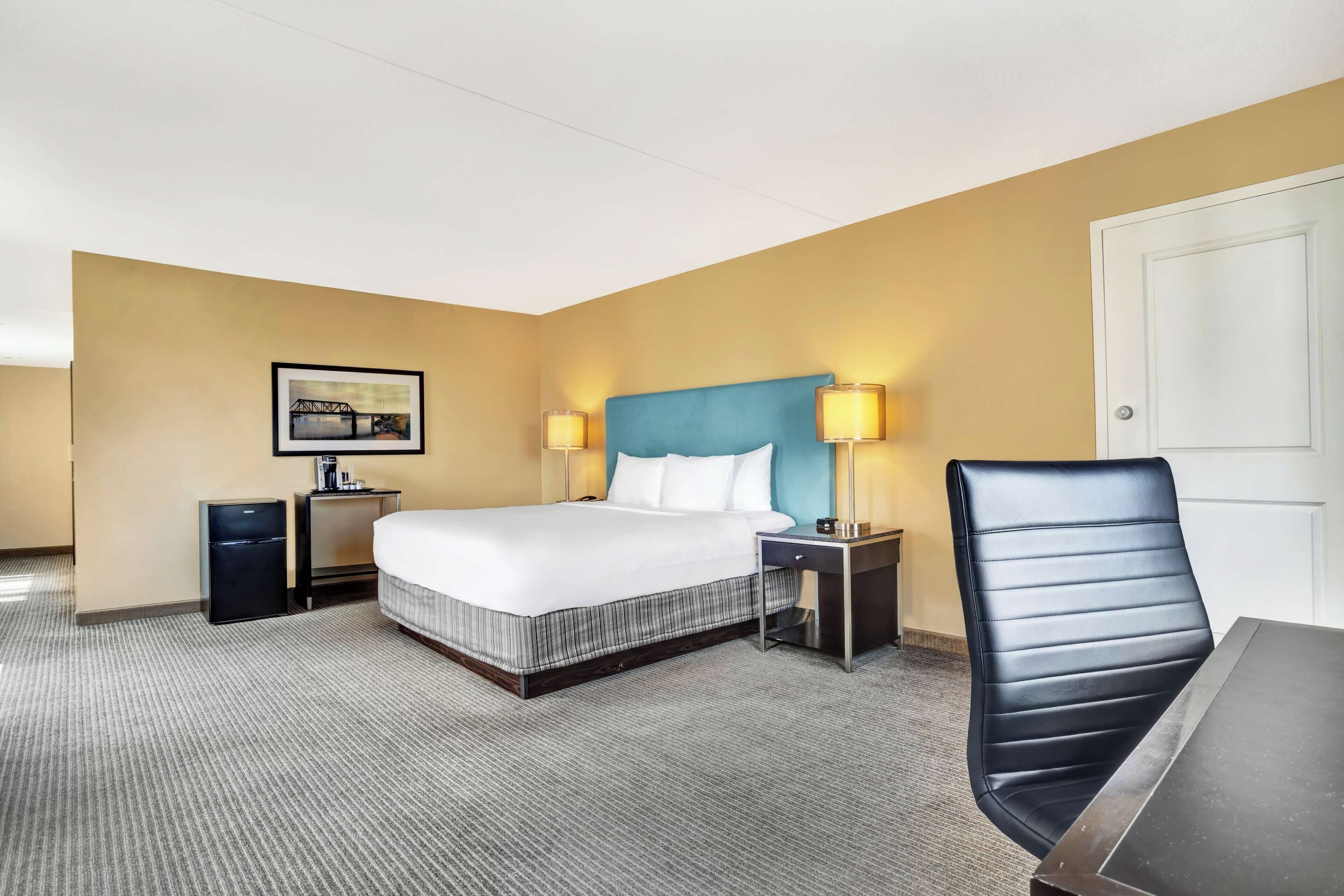 Hotelkamer met een bed, bureau, stoel, nachtkastje, lamp, foto aan de muur en een deur.