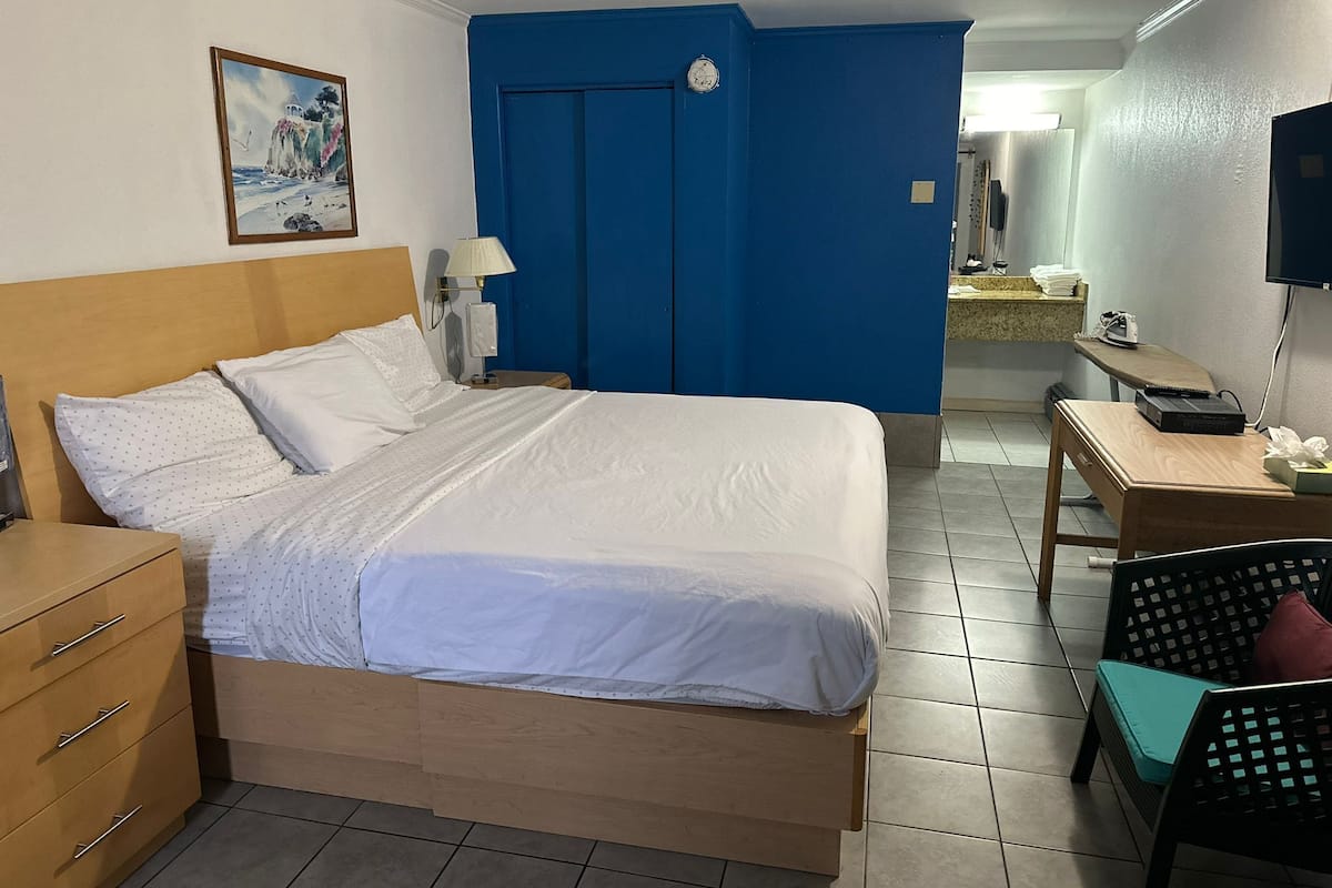A hotel room with a bed, a desk, a chair, a lamp, a clock, and a painting on the wall.