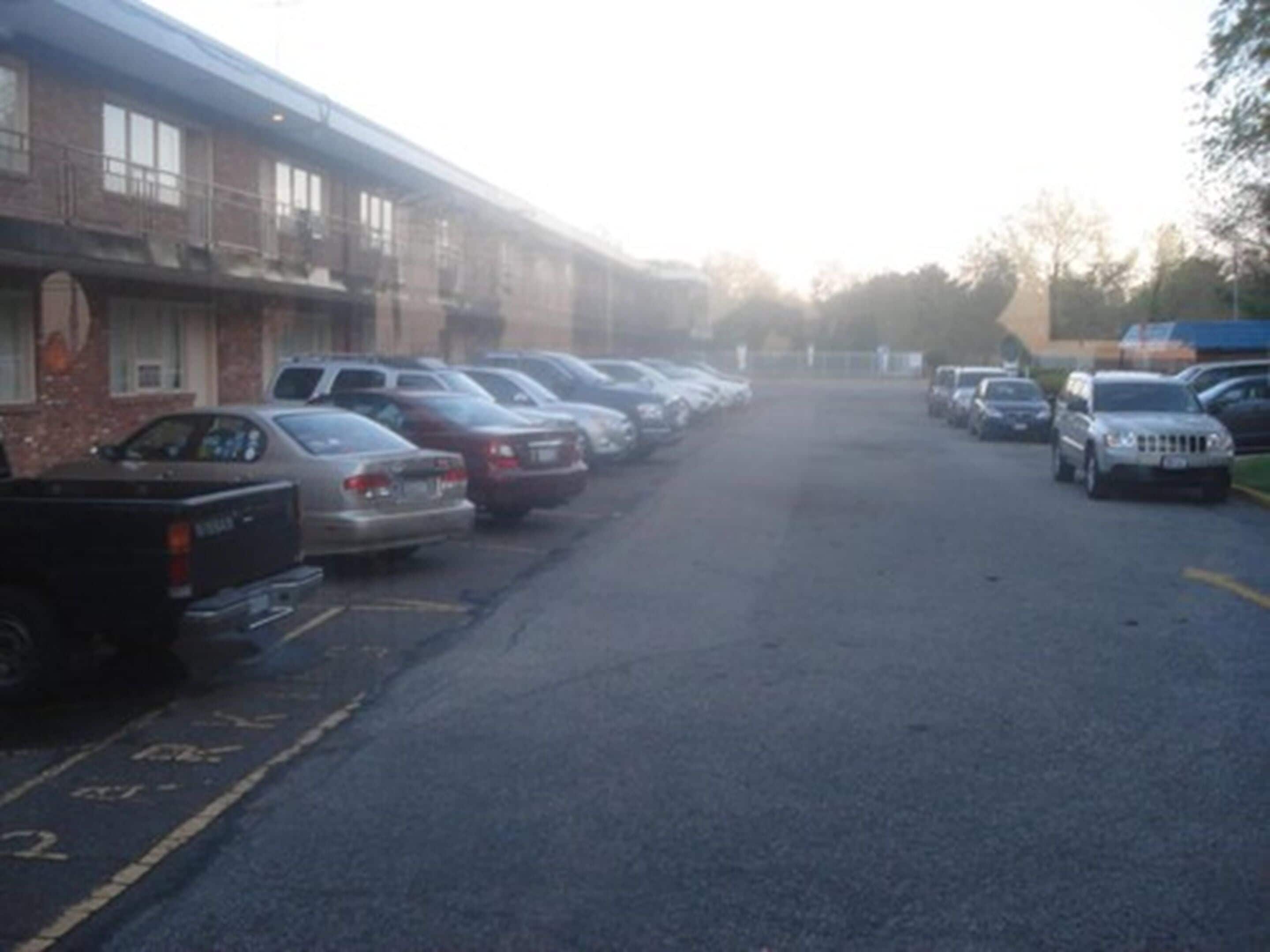 Un estacionamiento con varios coches aparcados en filas, un edificio de fachada de ladrillo y cielo despejado.